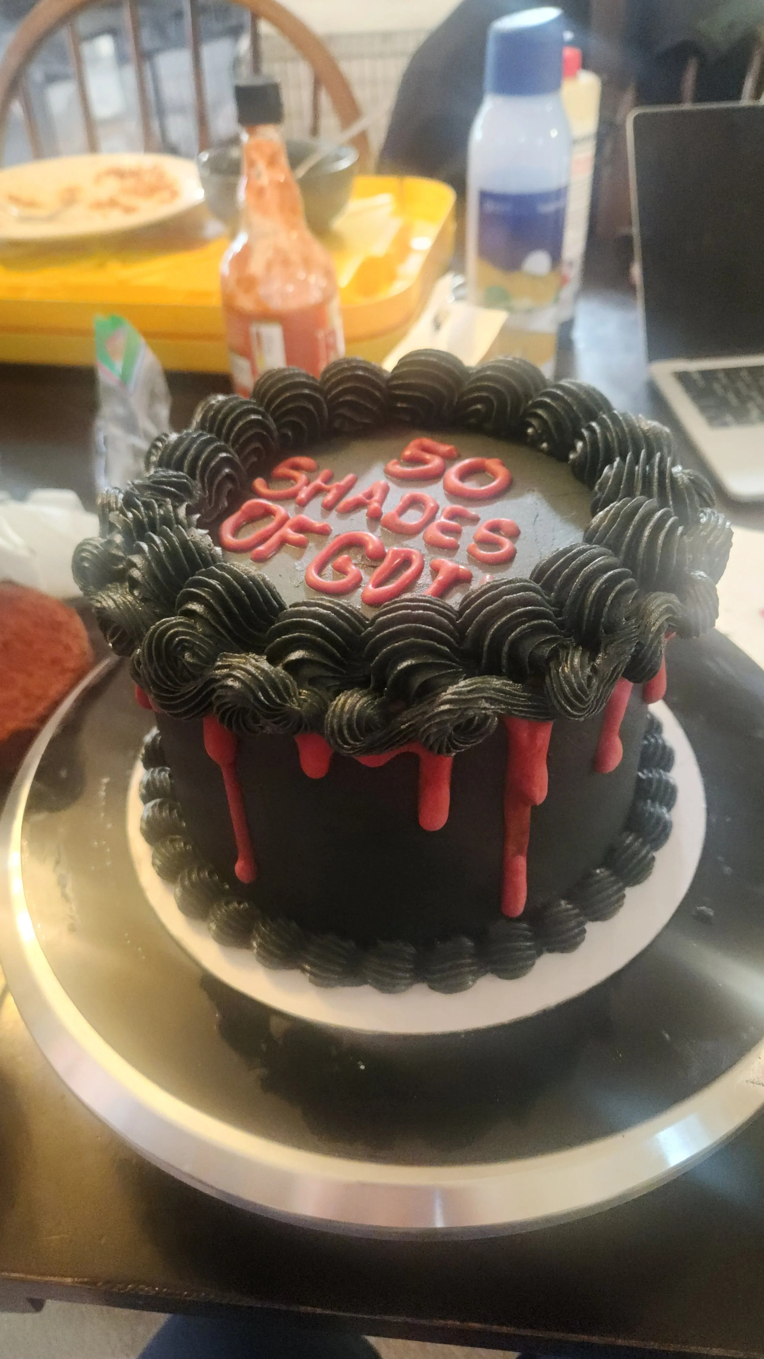 A birthday cake with black frosting, red drip decorations, and a message that reads '50 SHADES OF GDT' written in red icing, placed on a silver cake board.