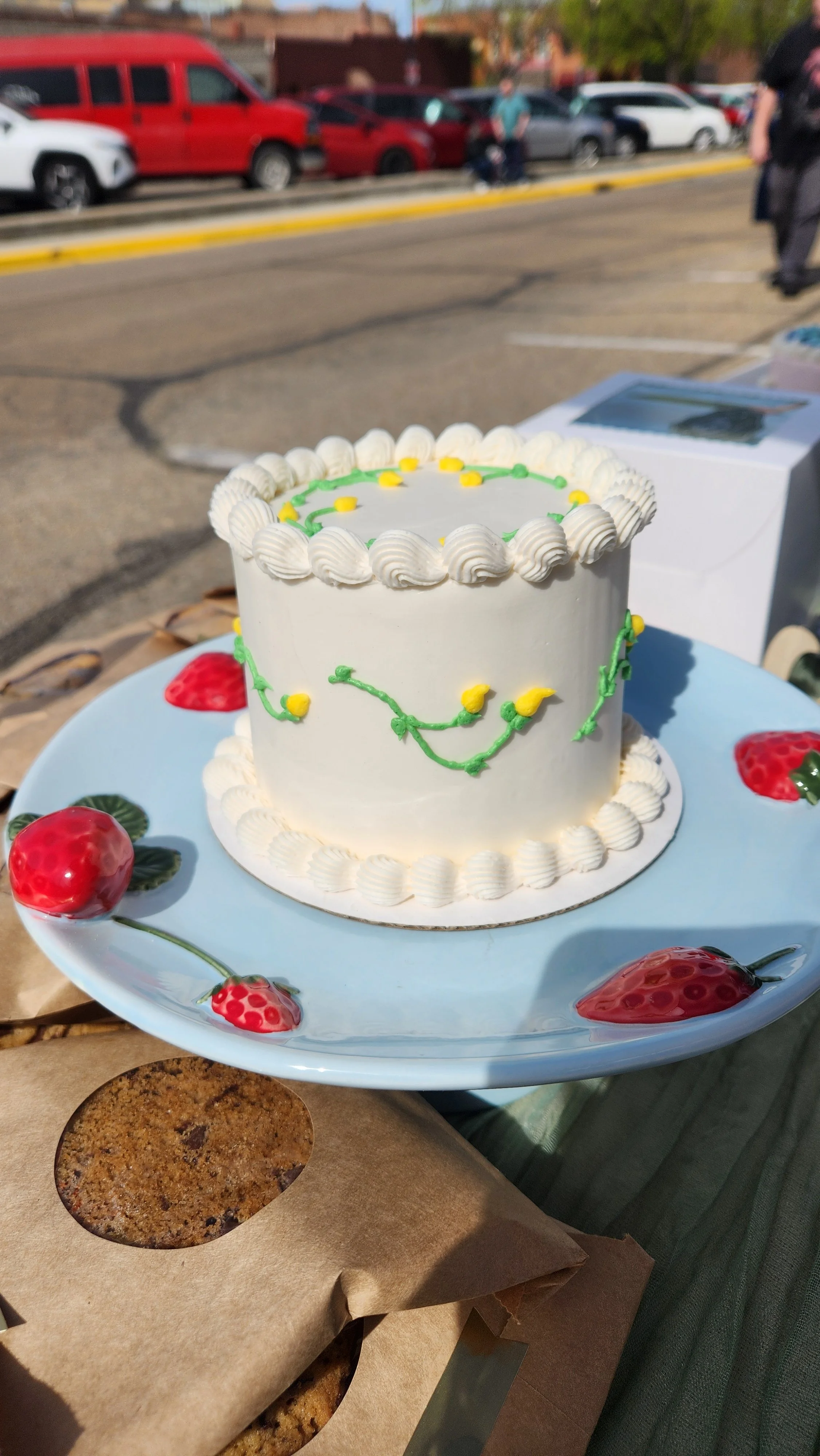 A white frosted cake decorated with green and yellow icing, sitting on a blue plate with strawberry-shaped decorations, outdoors with parking lot and cars in the background.
