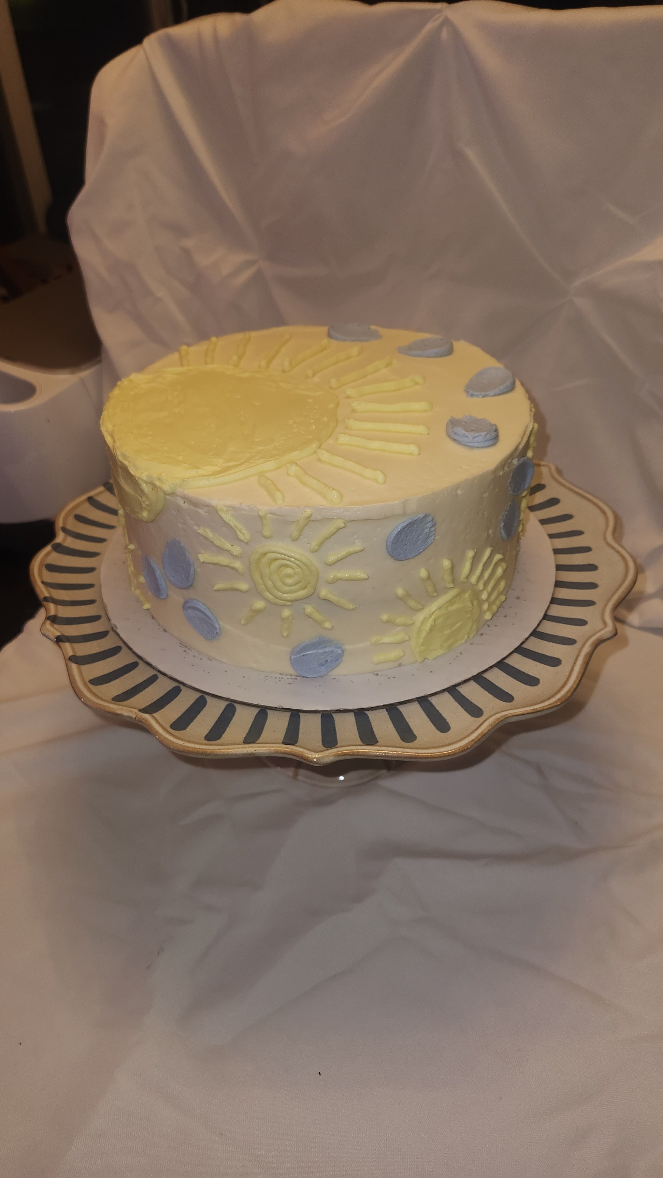 A round cake decorated with yellow and gray sun and moon designs, placed on a decorative cake stand with a black and gold rim, on a white tablecloth with a white fabric backdrop.