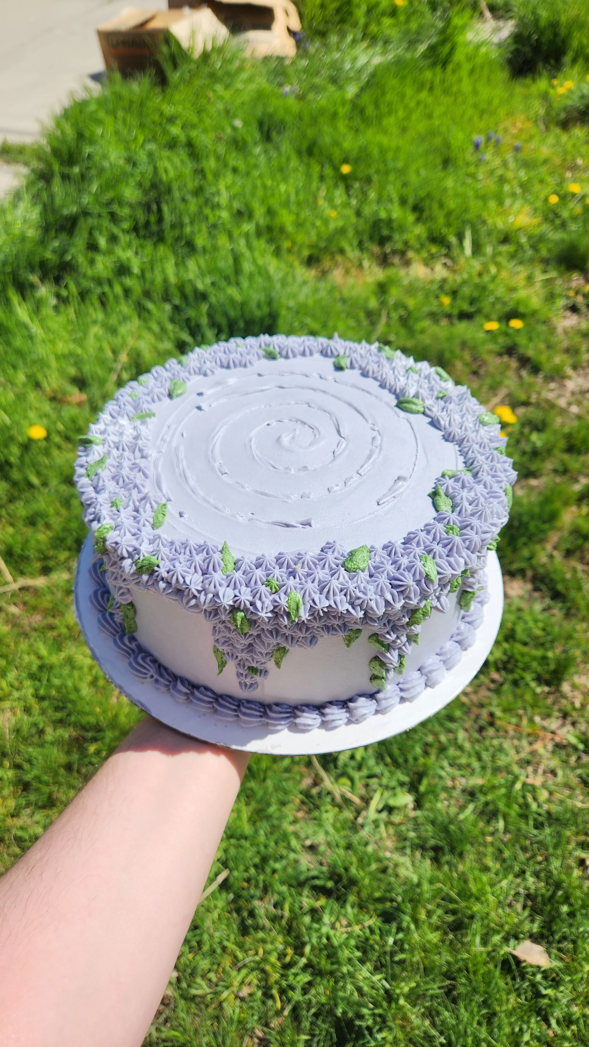 A white birthday cake decorated with purple and green icing flowers, being held outdoors over a grassy area.
