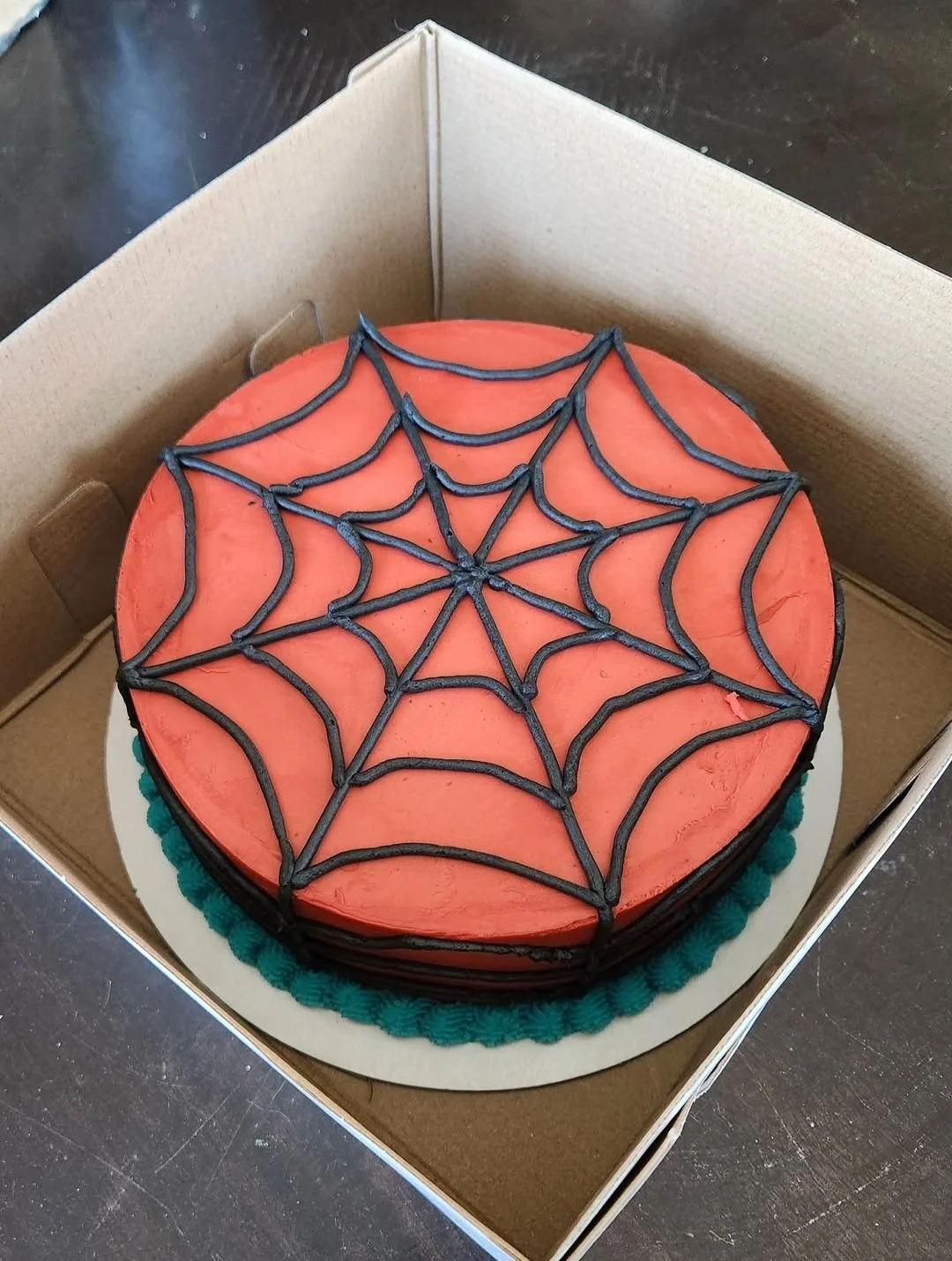 A round cake decorated with orange icing and a black spider web pattern on top, in a brown cardboard box.