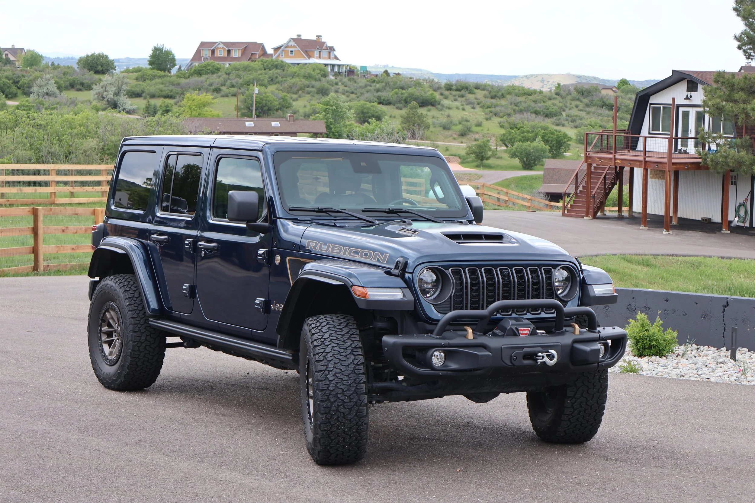 Black Jeep Rubicon parked on a paved surface with a wooden fence, green trees, houses and rolling hills in the background.