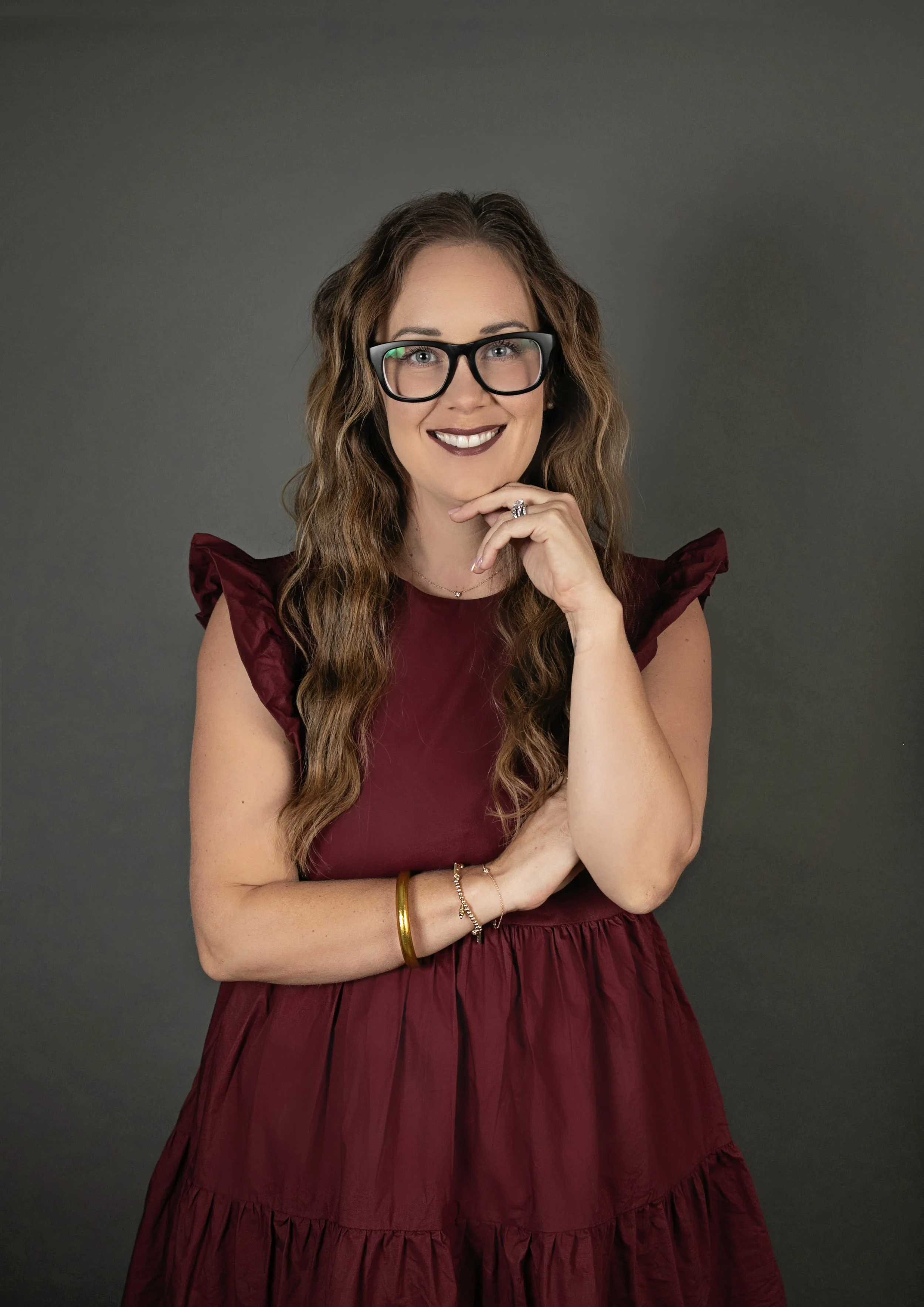 A woman with wavy brown hair and glasses, smiling with her hand near her chin, wearing a maroon dress with ruffled sleeves, standing against a gray background.