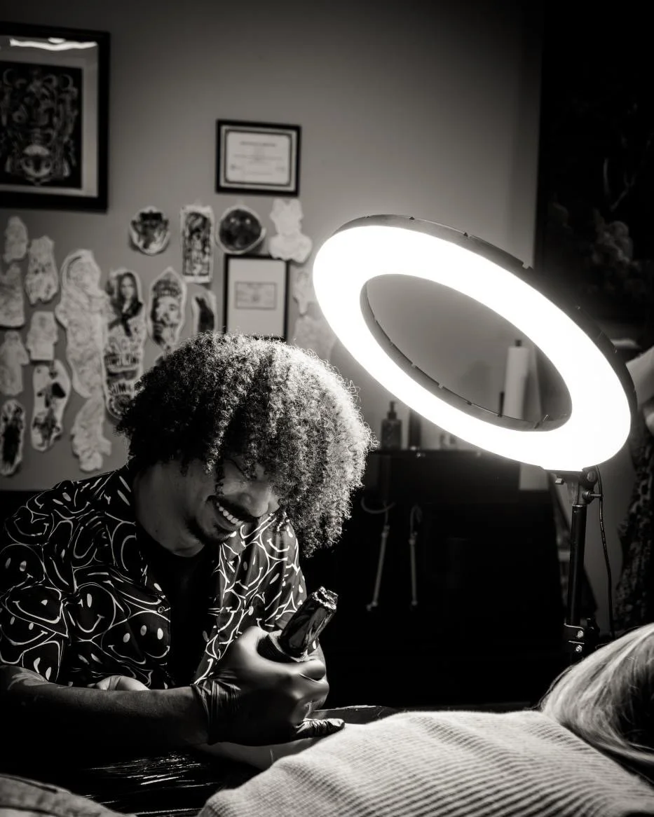 Tattoo artist with curly hair, wearing gloves, working on a client under a ring light in a tattoo shop.