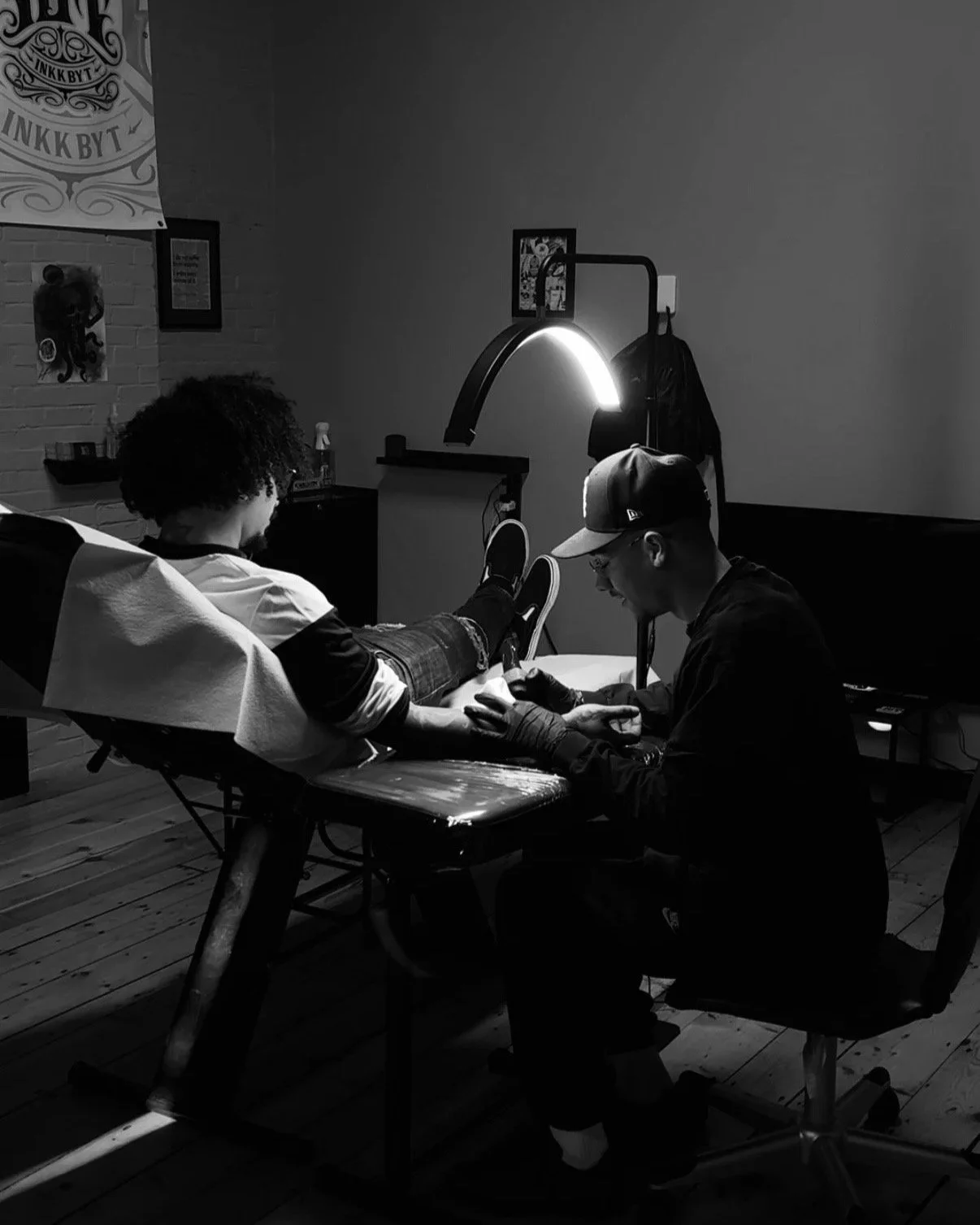 A person giving a tattoo to a client in a tattoo studio, both seated at a wooden table, with a lamp illuminating the area, and artwork hanging on the wall.