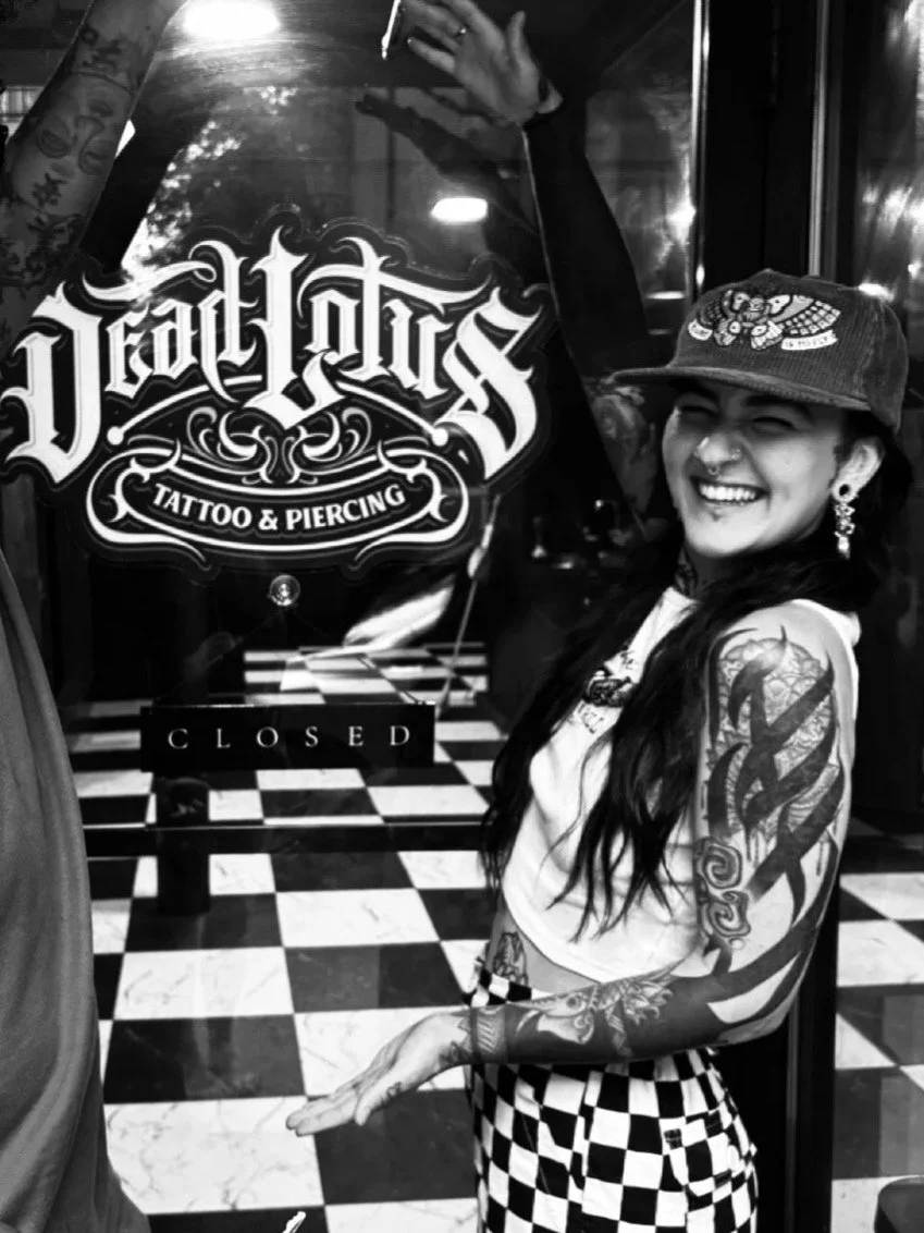 A smiling woman with tattoos on her arms, wearing a cap with a cartoon design, earrings, a crop top, and checkered pants, standing in front of a tattoo and piercing shop window with a "Closed" sign.