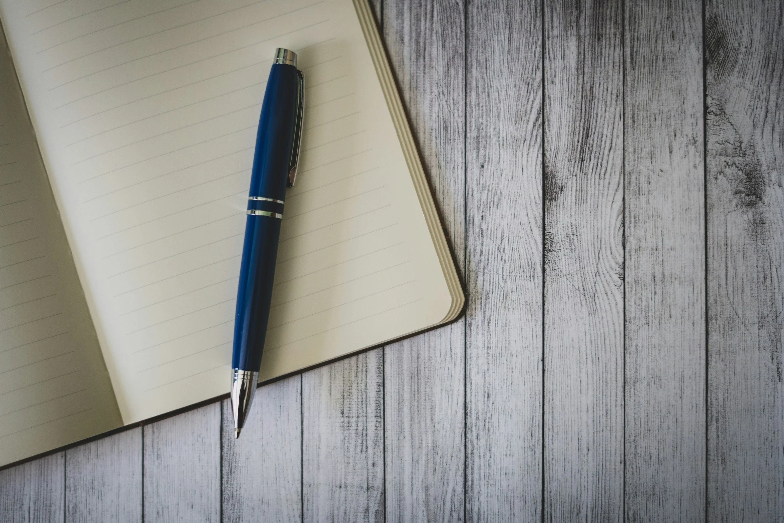 An open notebook with lined pages and a blue ballpoint pen resting on the left page, placed on a white wooden surface.