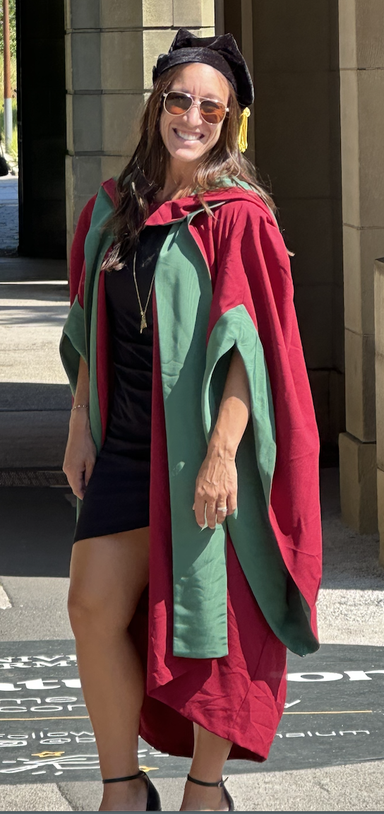 A woman in academic regalia, including a black hat with a yellow tassel, sunglasses, a black dress, and an academic gown with red and green panels, standing outside in sunlight.