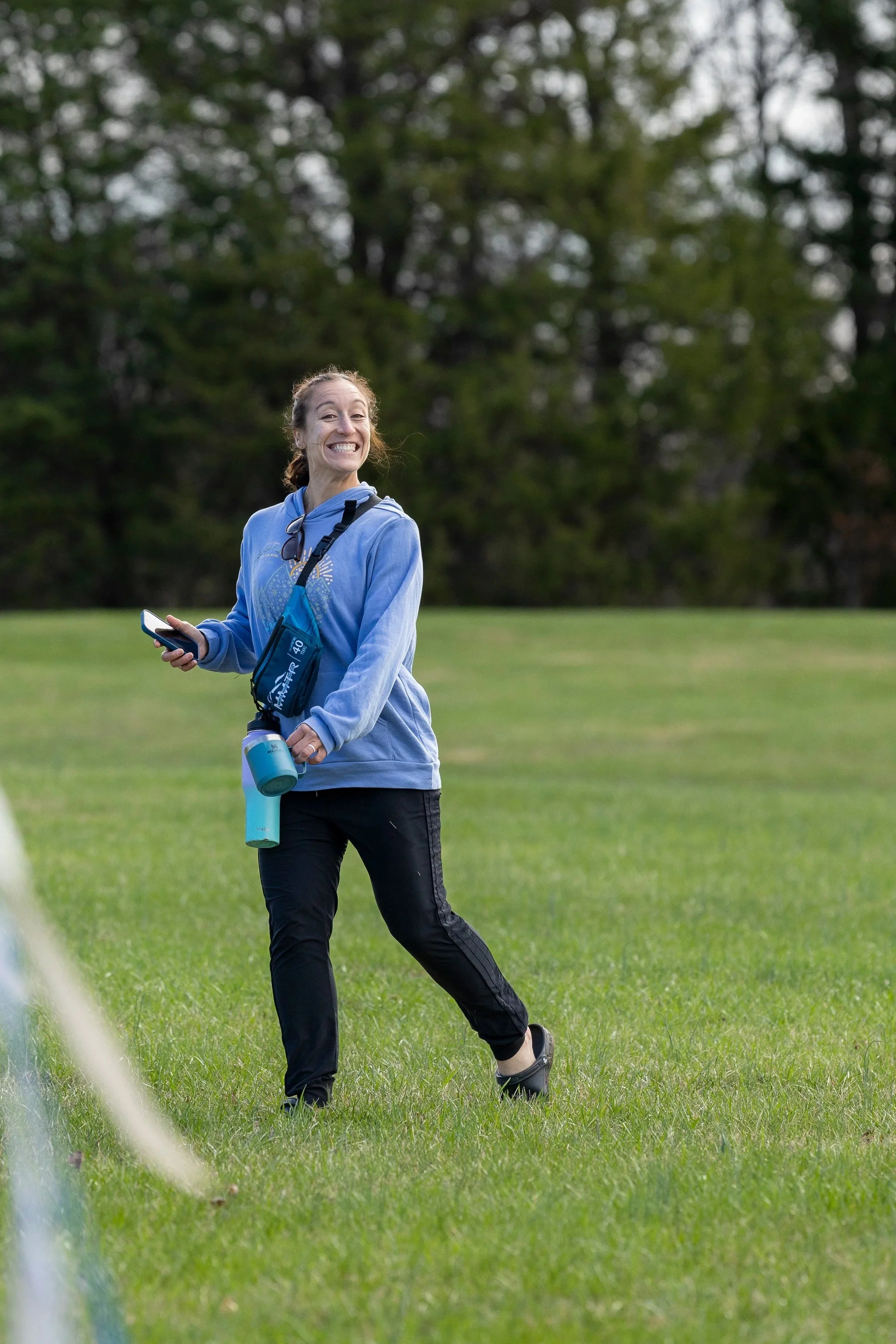 A woman outdoors on a grassy field, smiling and holding a phone and water bottle, dressed in athletic clothing with a blue hoodie and black pants, with trees in the background.