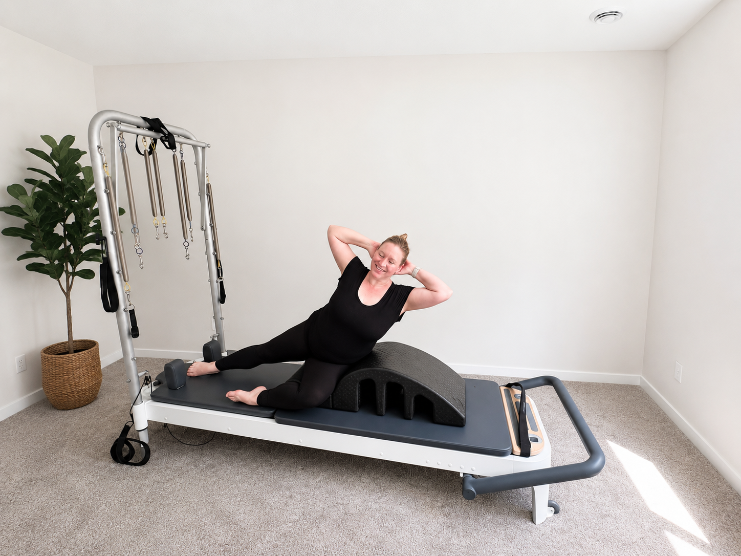A woman doing core exercises on a physical therapy or Pilates reformer machine, smiling with her hands behind her head, in a bright, minimalistic room with a potted plant.