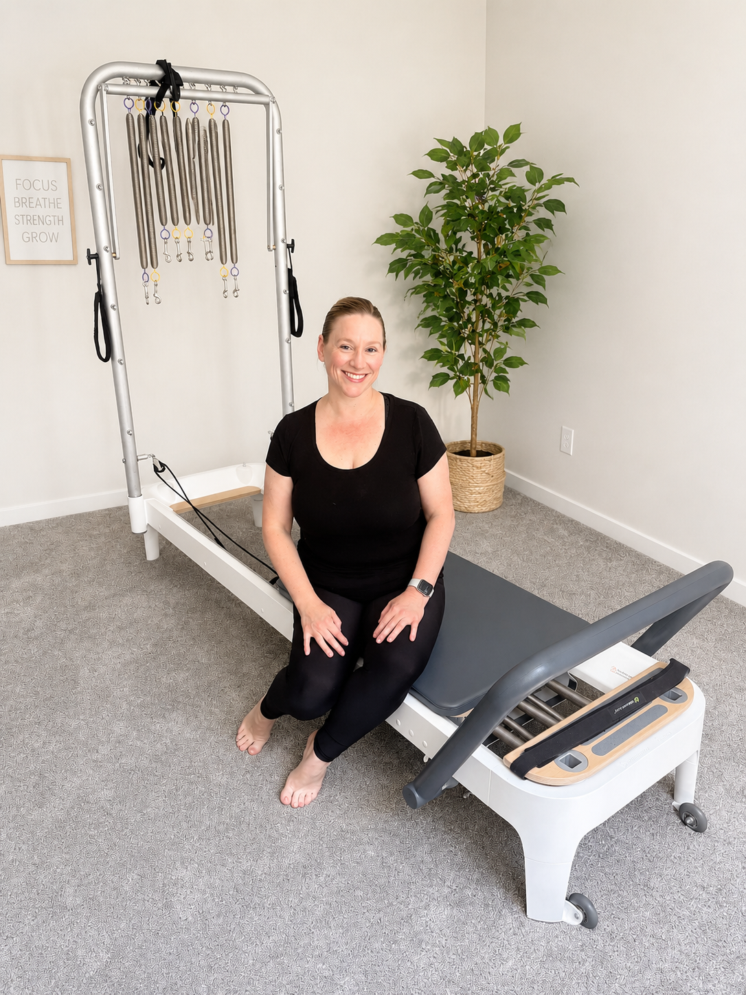 A woman sitting on a Pilates reformer machine in a therapy or fitness room, smiling, with a large potted plant behind her and a motivational sign on the wall that reads 'FOCUS BREATHE STRENGTH GROW'.