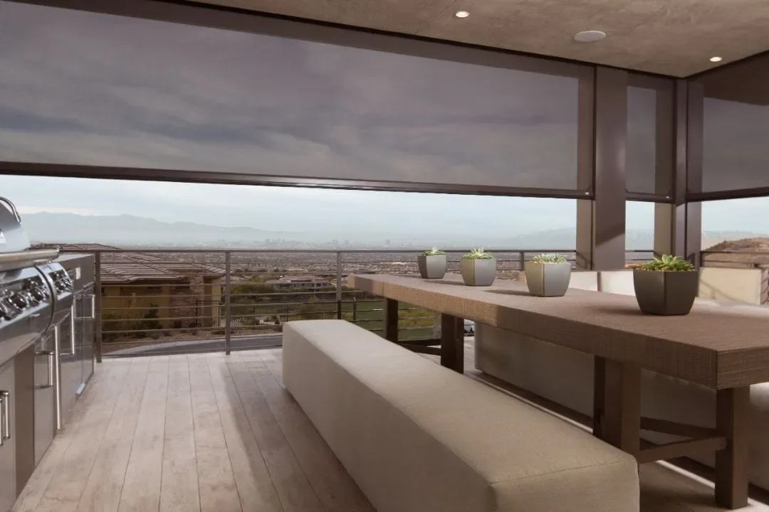 Modern outdoor balcony with a dining table and bench, potted plants, and a built-in grill, overlooking a city and mountain range in the distance.