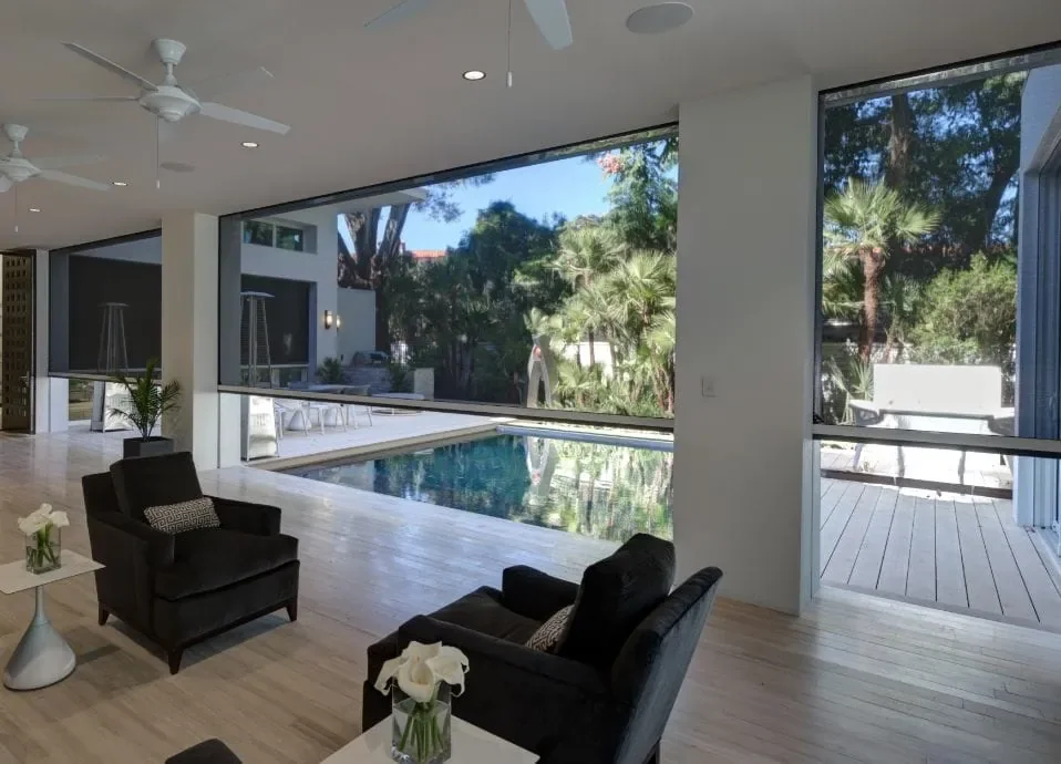 Modern indoor living area with large floor-to-ceiling windows overlooking a backyard with a pool, outdoor seating, and lush trees.