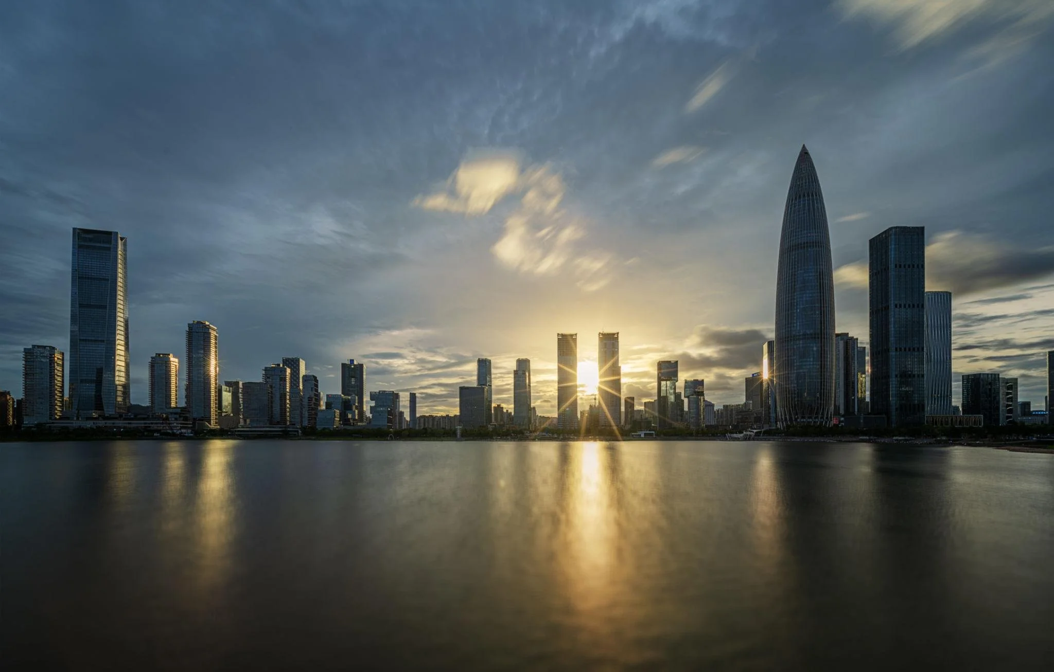 City skyline with tall skyscrapers during sunset, reflecting on water in the foreground.