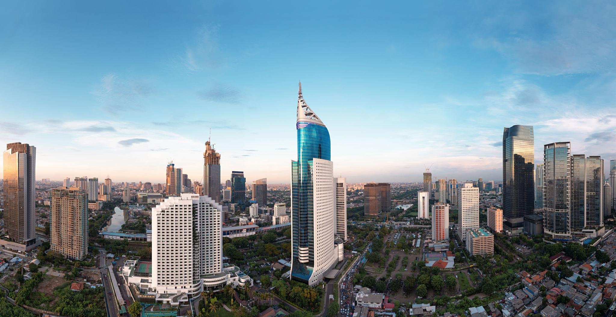 A city skyline featuring modern skyscrapers under a clear blue sky with some clouds. Notable tall buildings include a blue glass tower with a pointed top.