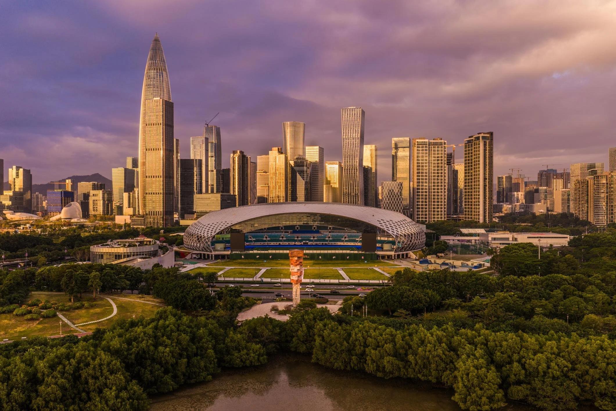 A city skyline during sunset featuring tall modern skyscrapers, a large stadium with a white roof, and lush green trees along a river in the foreground.