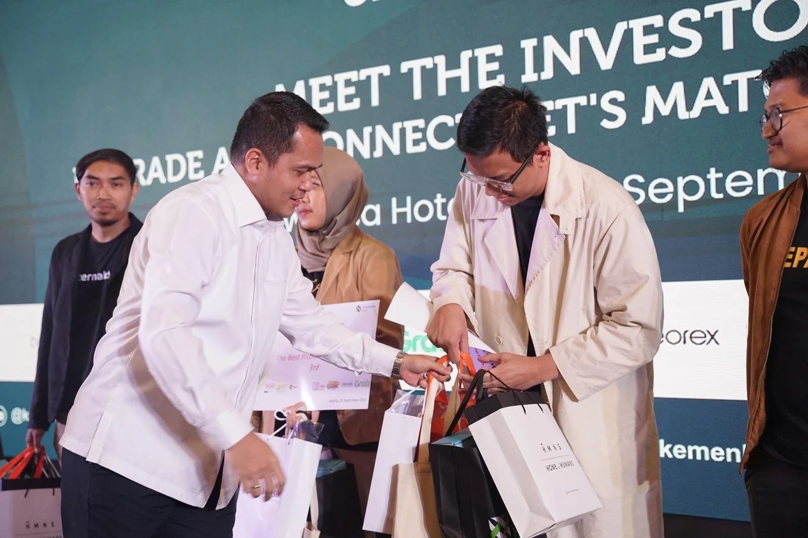 People exchanging gifts at a conference or event with a large screen displaying text in the background.