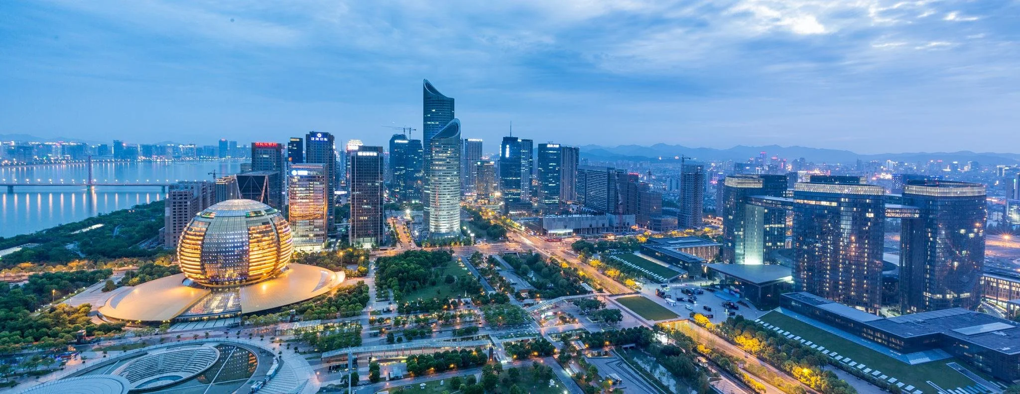 Skyline of a modern city at dusk with illuminated skyscrapers, a river on the left, and a park with green trees in the foreground.
