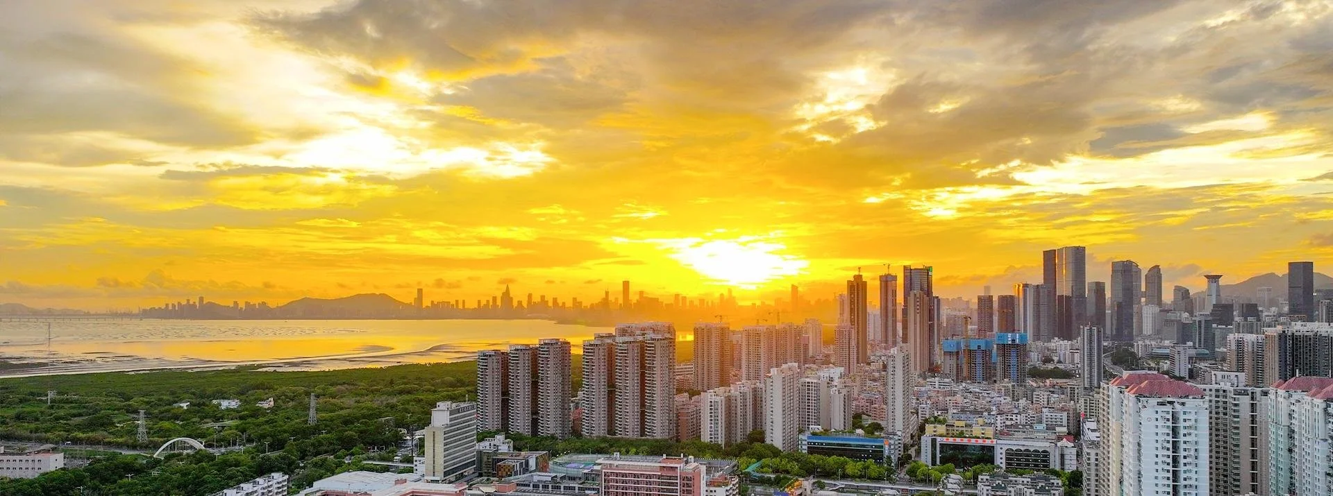 Sunset over a city skyline with high-rise buildings, a body of water, and mountains in the background, with a yellow and orange sky.