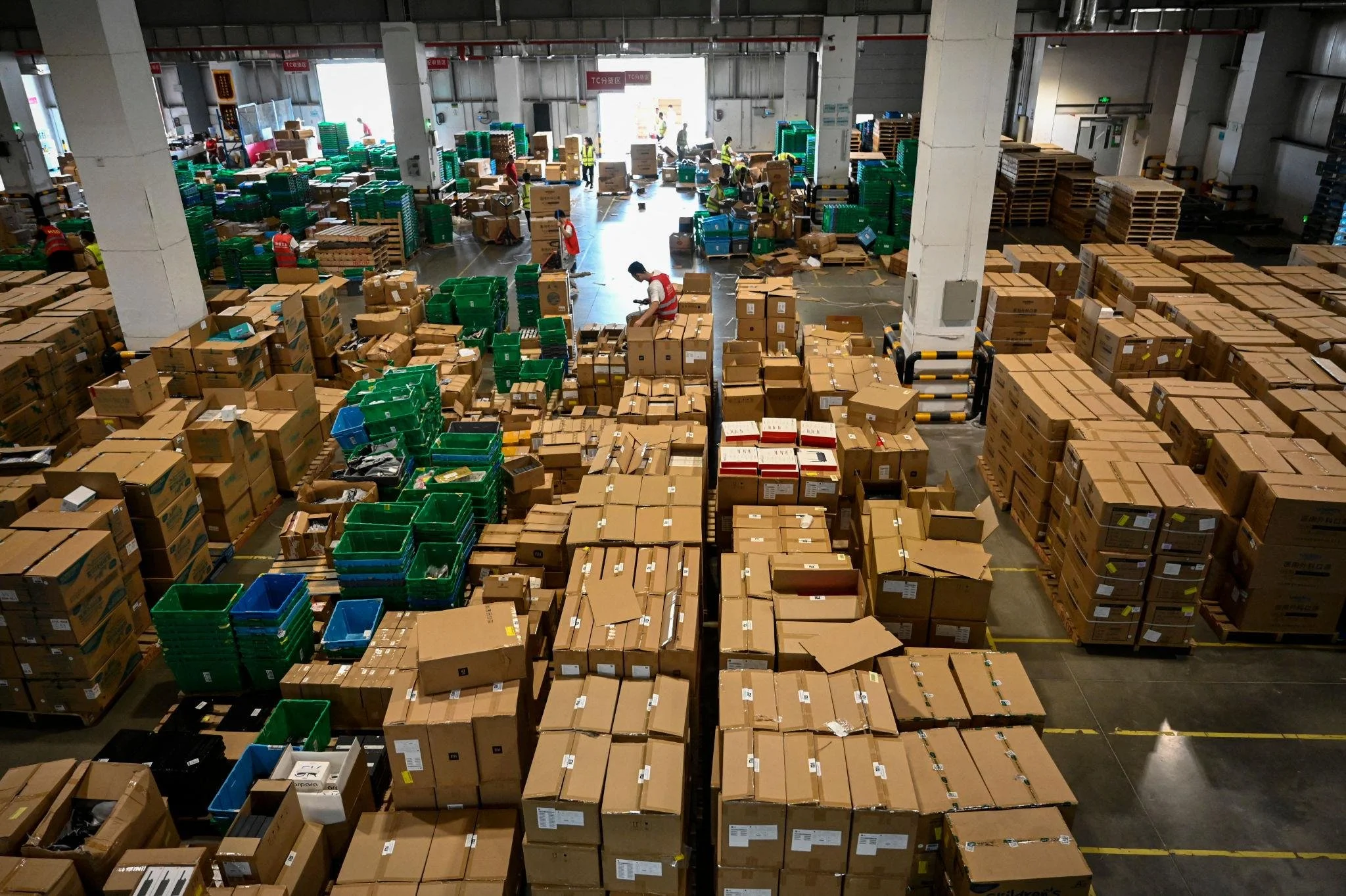 Warehouse interior filled with stacks of cardboard boxes and plastic bins, with workers wearing safety vests handling packages.