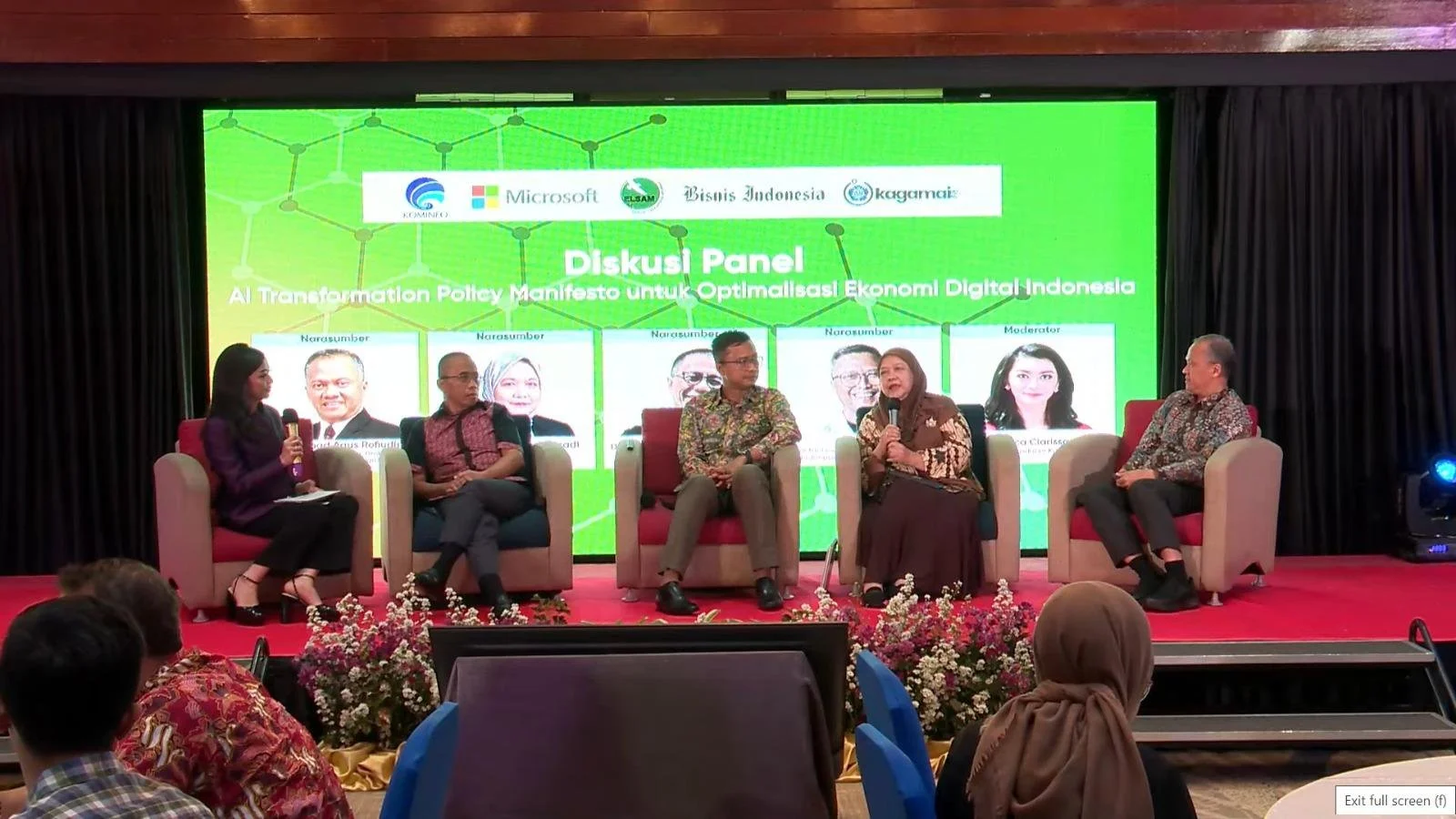 A panel discussion at a conference with five people on stage. Four are seated in chairs, one is standing. Background shows a bright green screen with logos and text about a discussion panel on digital economy and AI transformation. Audience members a