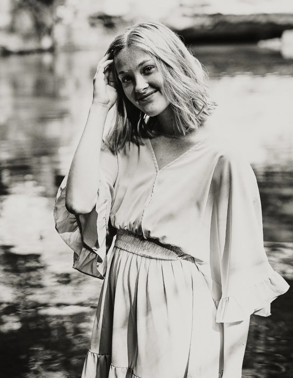A woman with shoulder-length wavy hair smiling, standing outdoors near water, wearing a light-colored dress with ruffled sleeves.