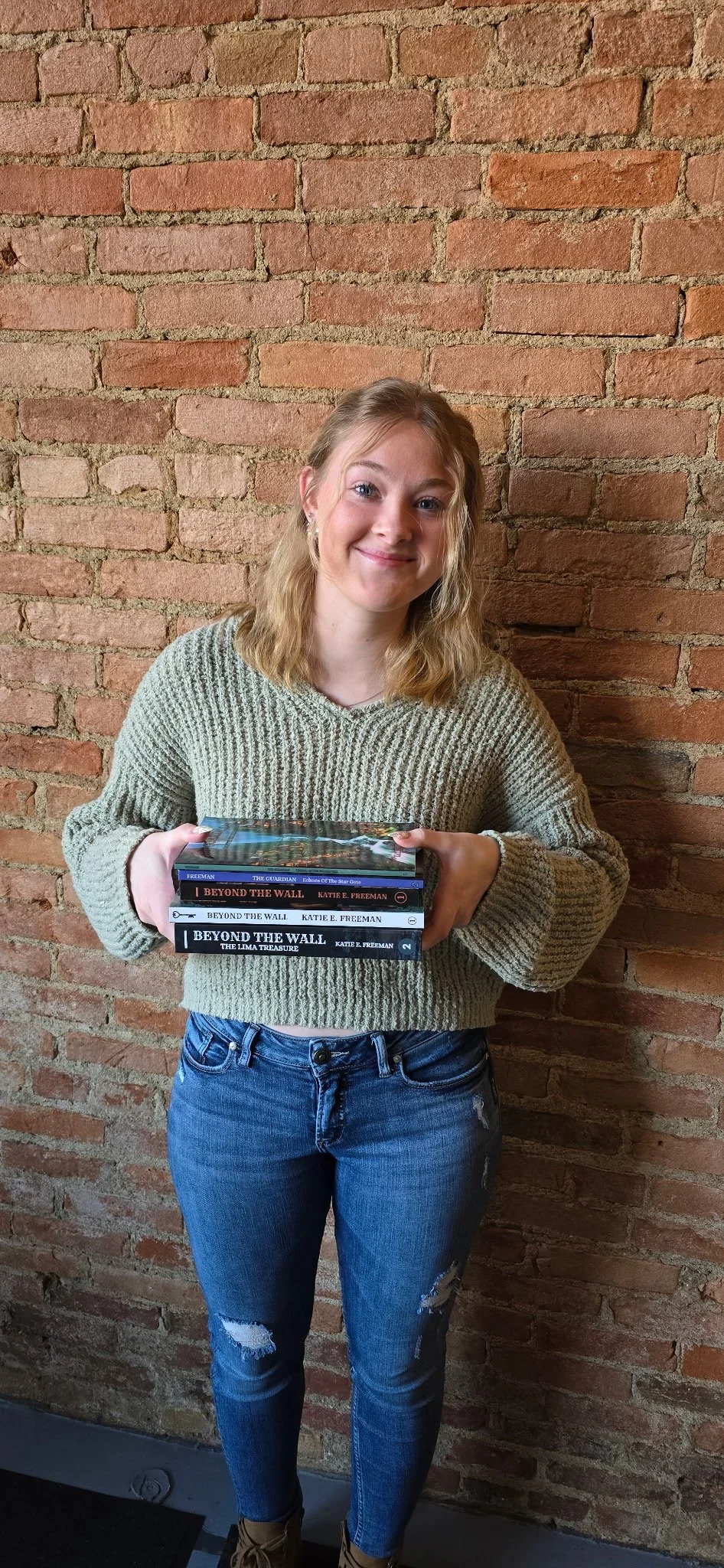 A young woman with blonde hair, wearing a beige knitted sweater and blue ripped jeans, is standing against a brick wall. She is smiling and holding a stack of five books titled 'Beyond The Wall' by Katie E. Freeman and other titles.