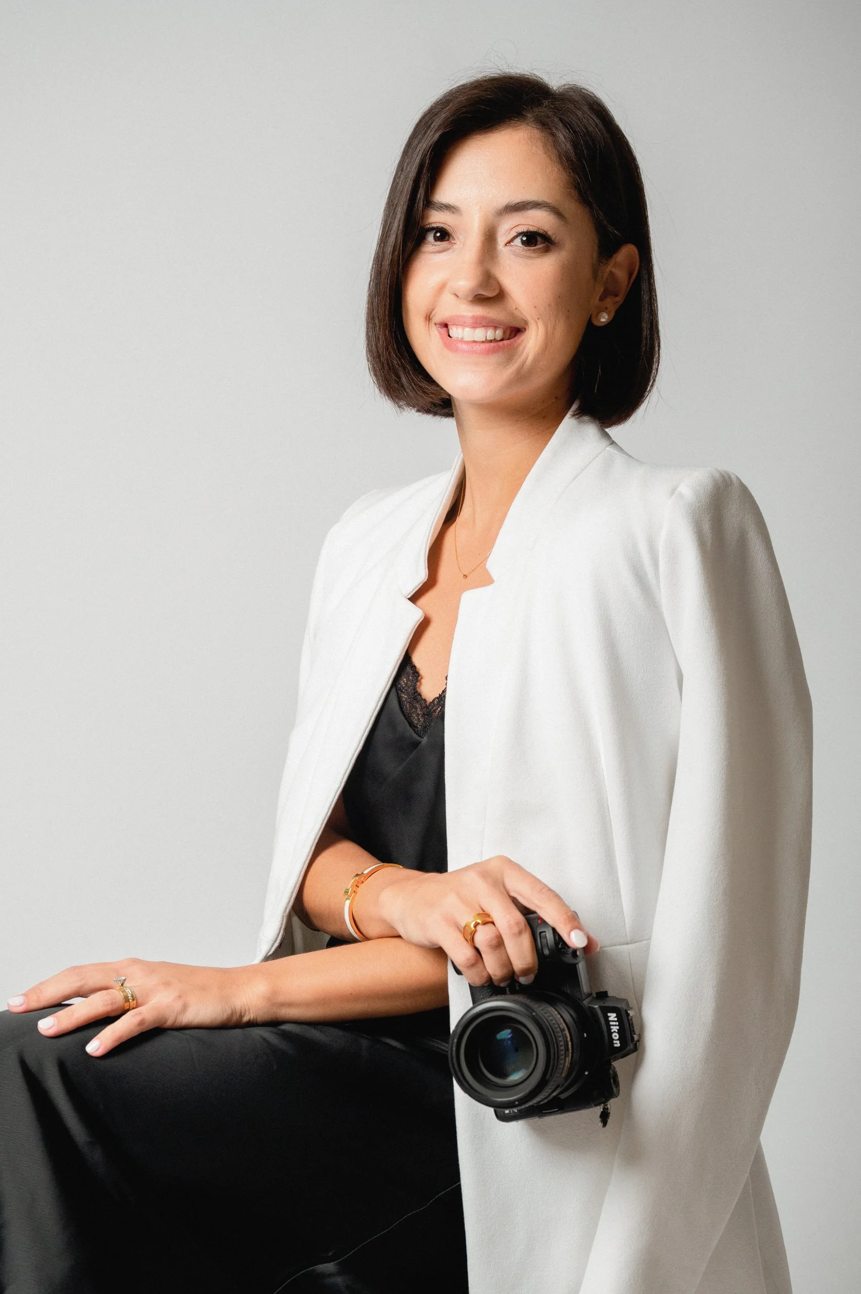 Darllane with shoulder-length dark hair, smiling, holding a Nikon camera, wearing a white blazer, jewelry, and black clothing underneath, sitting in front of a plain light gray background.