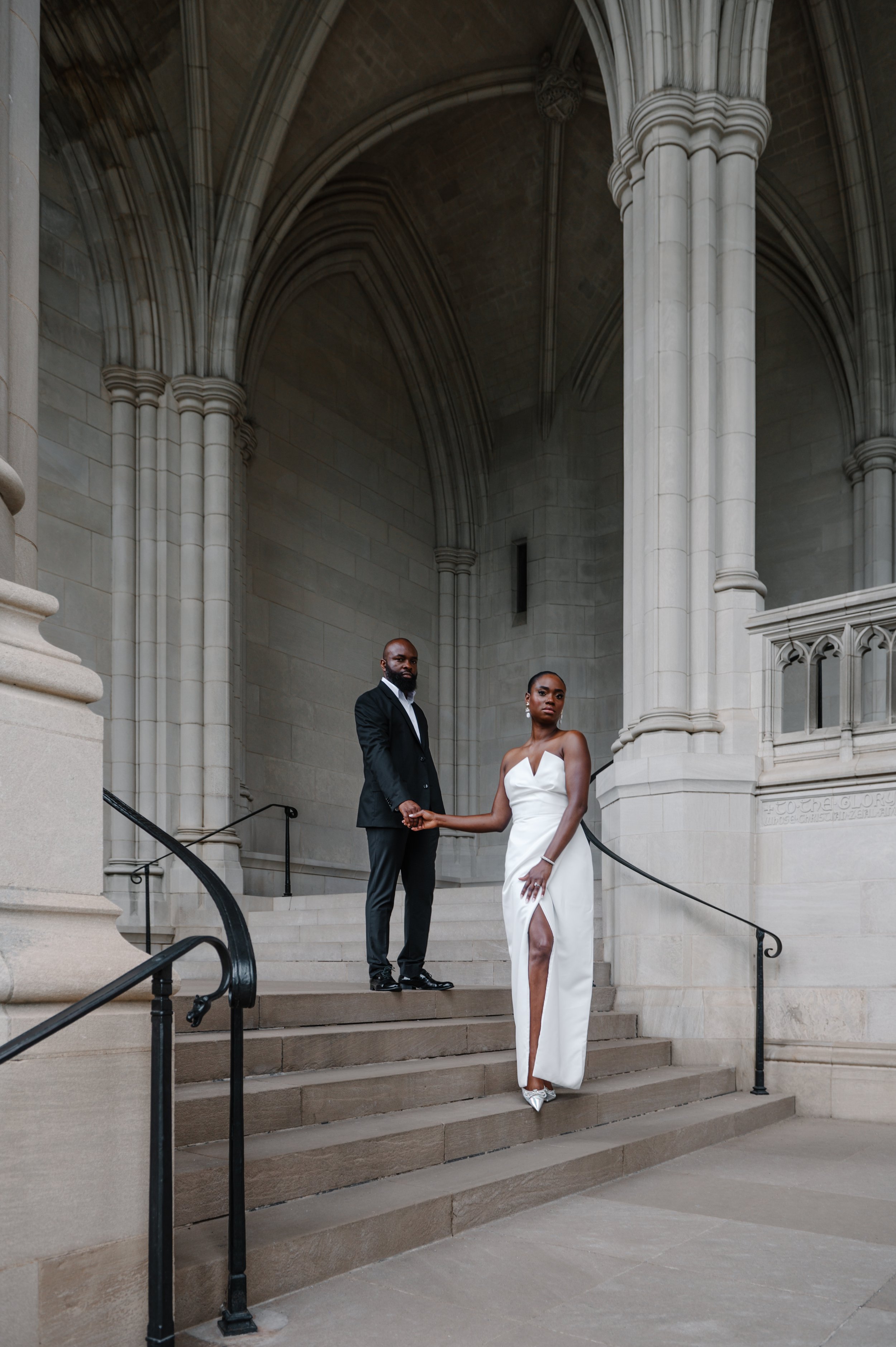 A man and a woman standing on the steps of the historic Washington National Cathedral with gothic architecture. The woman is wearing a white strapless dress with a slit, and silver heels, while the man is dressed in a black suit.