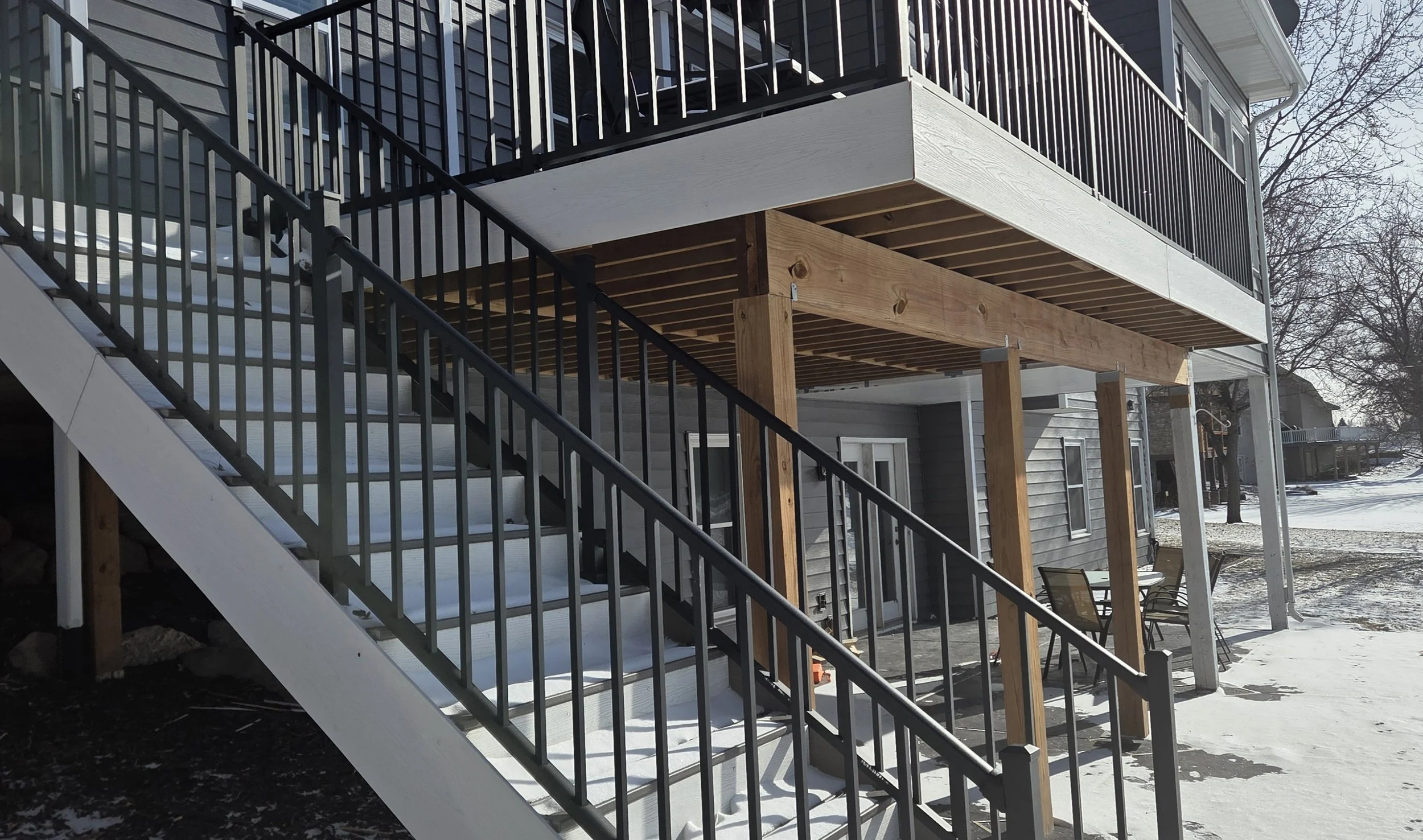 View of a house with a staircase leading to a second-floor balcony, construction work on the balcony with exposed wood beams, and snow covering the ground.