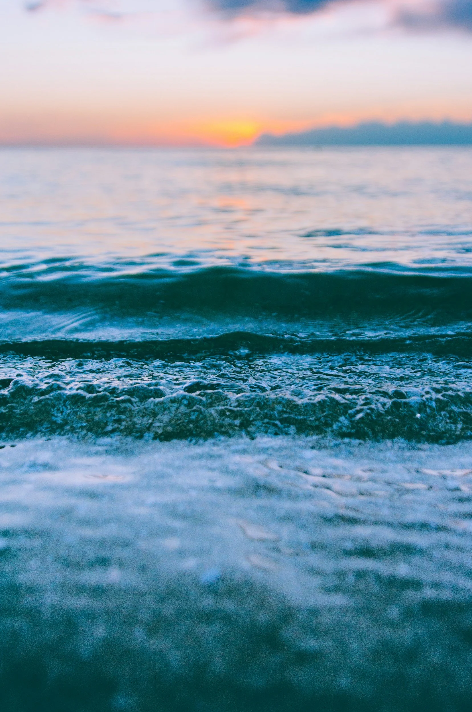 A blurry photo of the ocean during sunset with gentle waves in the foreground and a colorful sky with clouds in the background.
