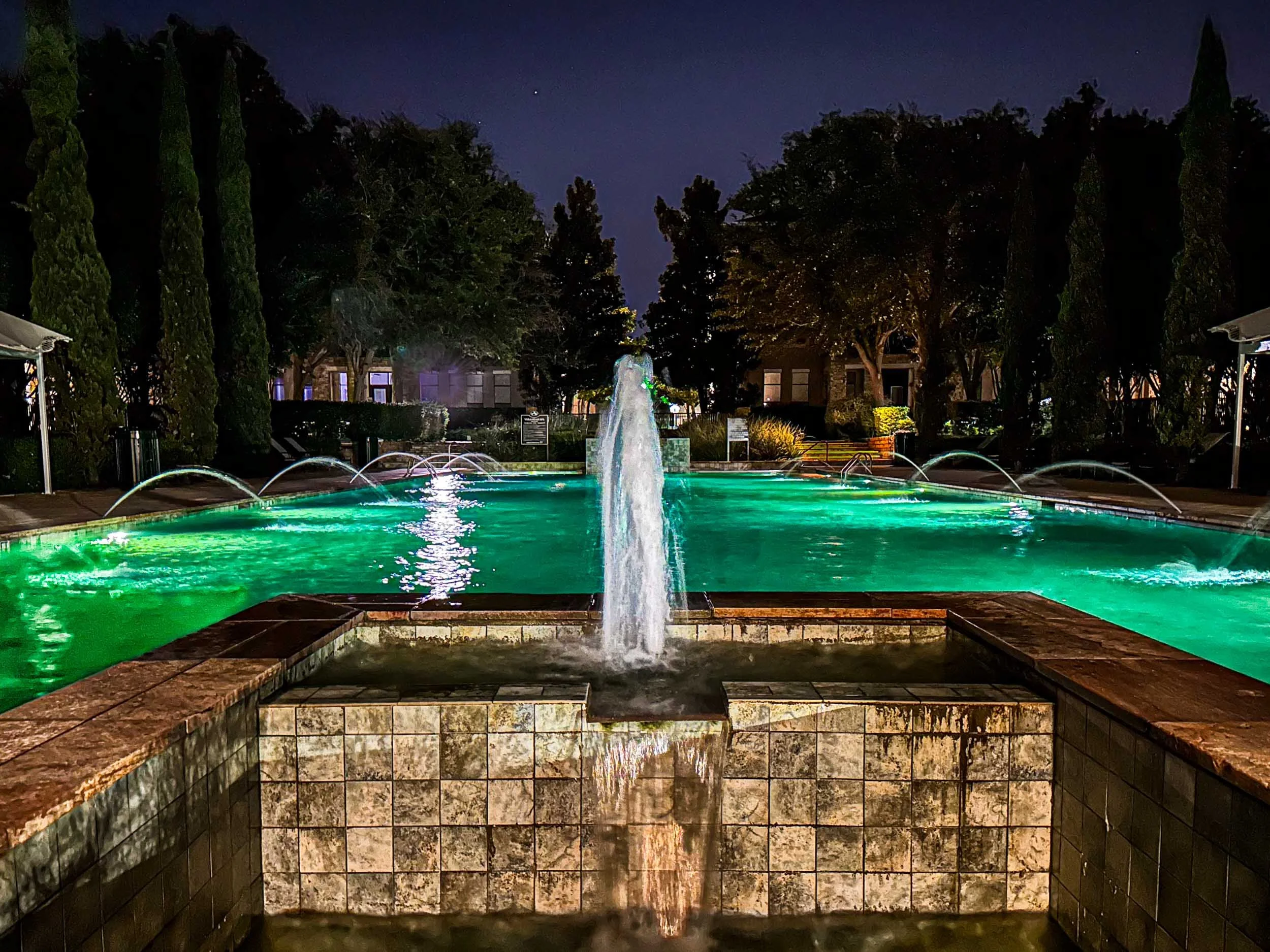 A community pool in Austin Ranch (Plano TX). The pool is illuminated at night with streams of water along each side and a decorative fountain in the foreground.