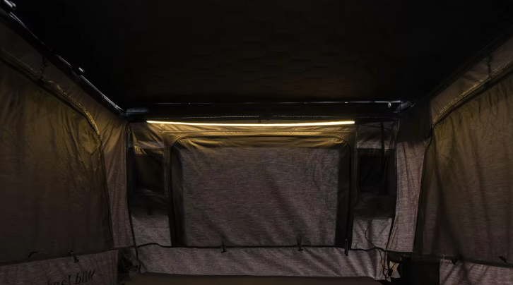 The interior of a small pop-up camper or tent with fabric walls and a sleeping area, dimly lit with a small strip of light at the top.