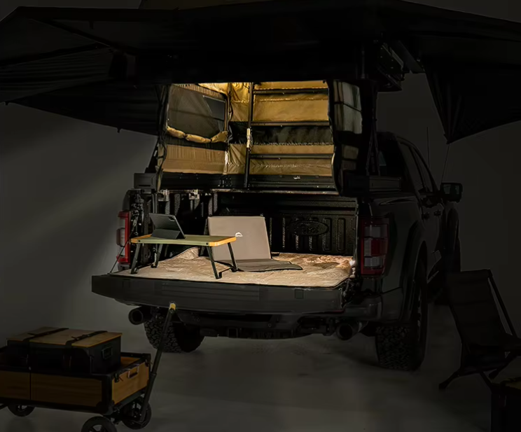 The bed of a pickup truck with a mounted sleeping tent, a small desk, and a chair inside the tent. There is a small cart with equipment nearby.