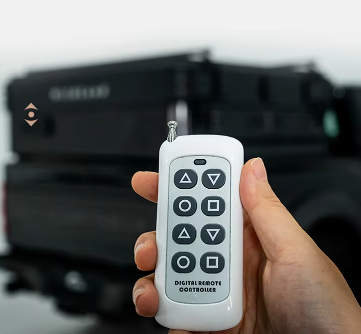 A hand holding a small white remote control with gray buttons, in front of a black machine or device in the background.