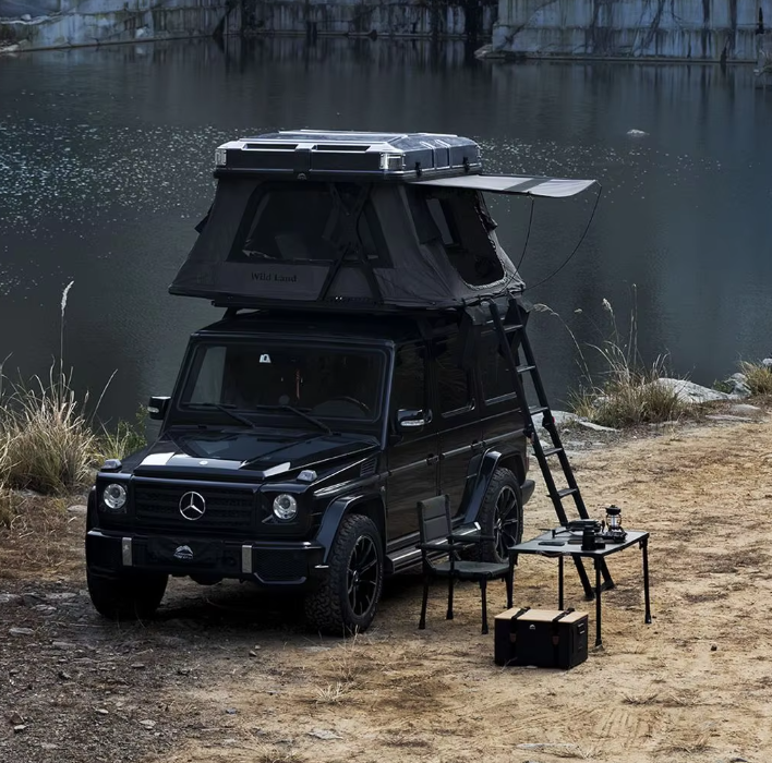 A black Mercedes-Benz G-Class vehicle with a rooftop tent set up by a body of water. There is a small table with chairs and equipment setup next to the vehicle, indicating an outdoor camping scene.