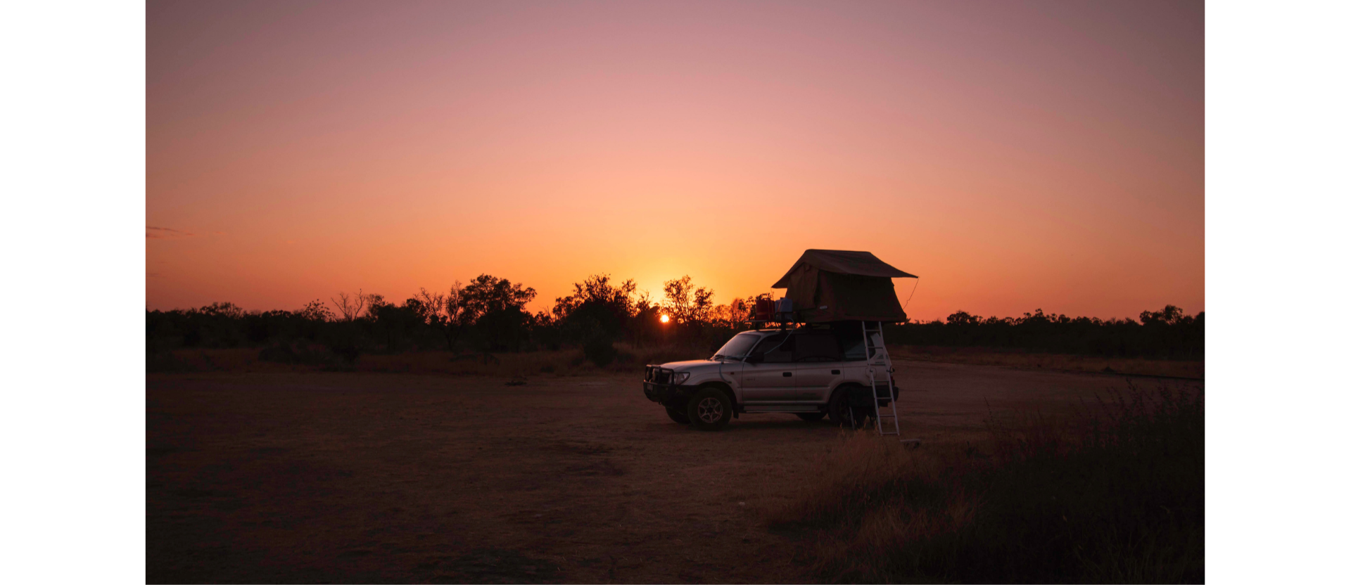 Sunset scene with a vehicle that has a rooftop tent and ladder set up in an open landscape with trees in the background.