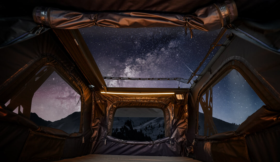 Inside of a tent with an open roof showing a starry night sky and the Milky Way galaxy, with mountains in the distance.