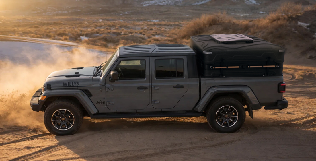 A gray Jeep Gladiator pickup truck driving on a dirt trail in a desert landscape, kicking up dust, with a large black cargo box mounted on the truck bed.