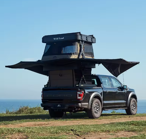A black pickup truck with a rooftop camper and an awning extended on one side, parked on grassy ground near the coast.