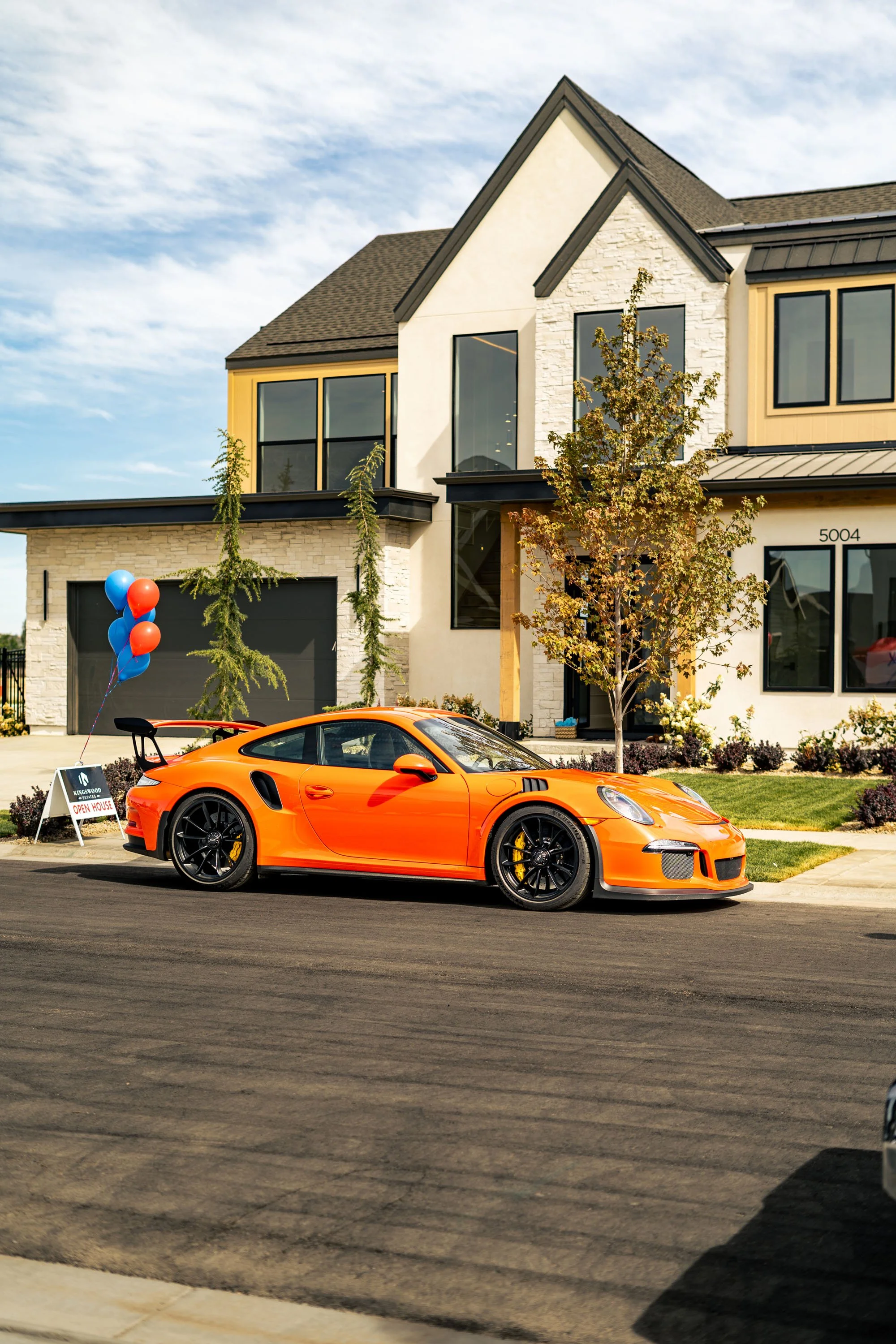 A bright orange sports car parked in front of a modern house with a gray roof, white walls, and large windows. There are balloons and an 'Open House' sign near the car.