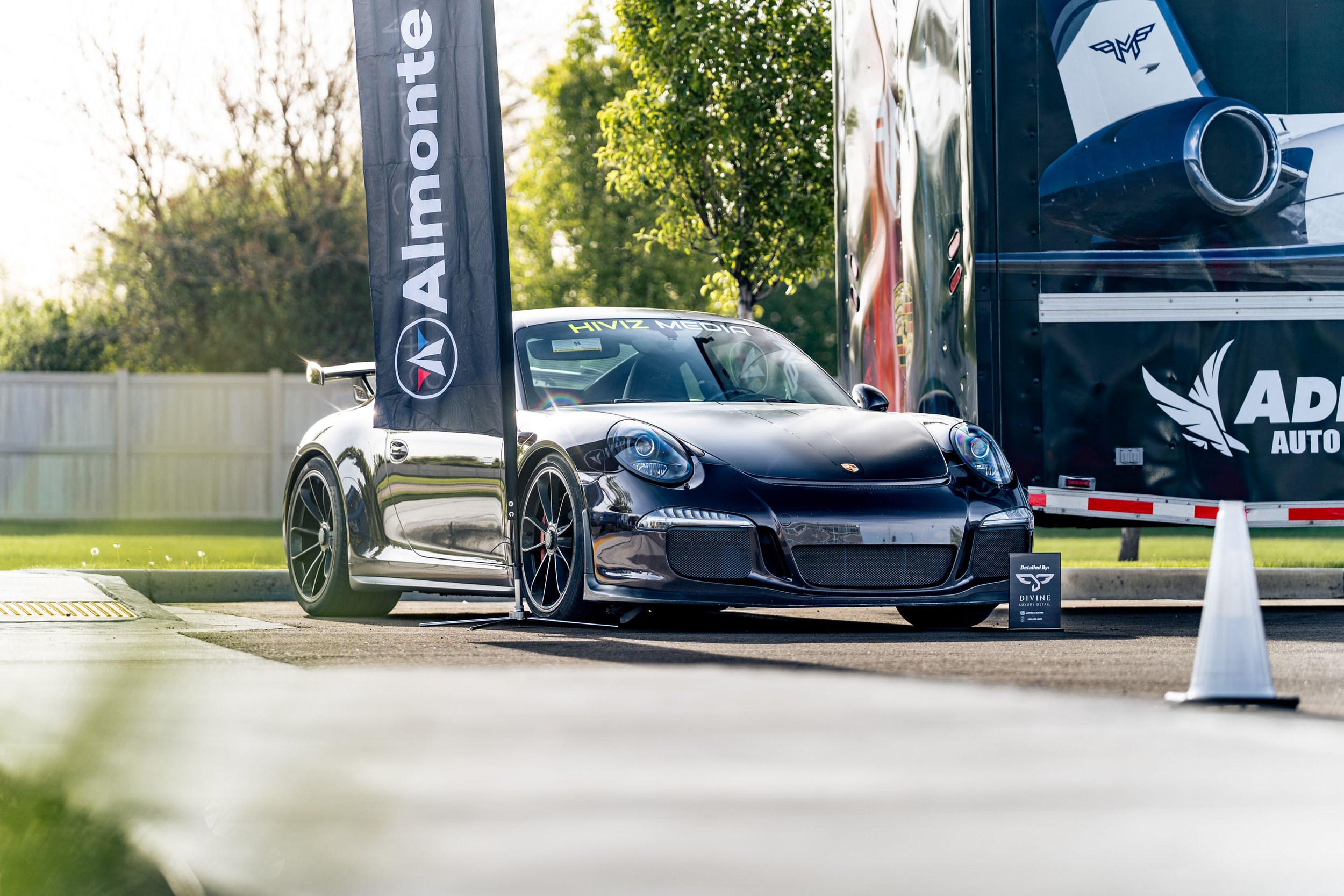 A black sports car with a Porsche logo is parked outdoors next to a black flag with white text reading 'Aston' and a logo. The background features green trees and a white fence, with a large black truck trailer behind the car. The car has tinted wind