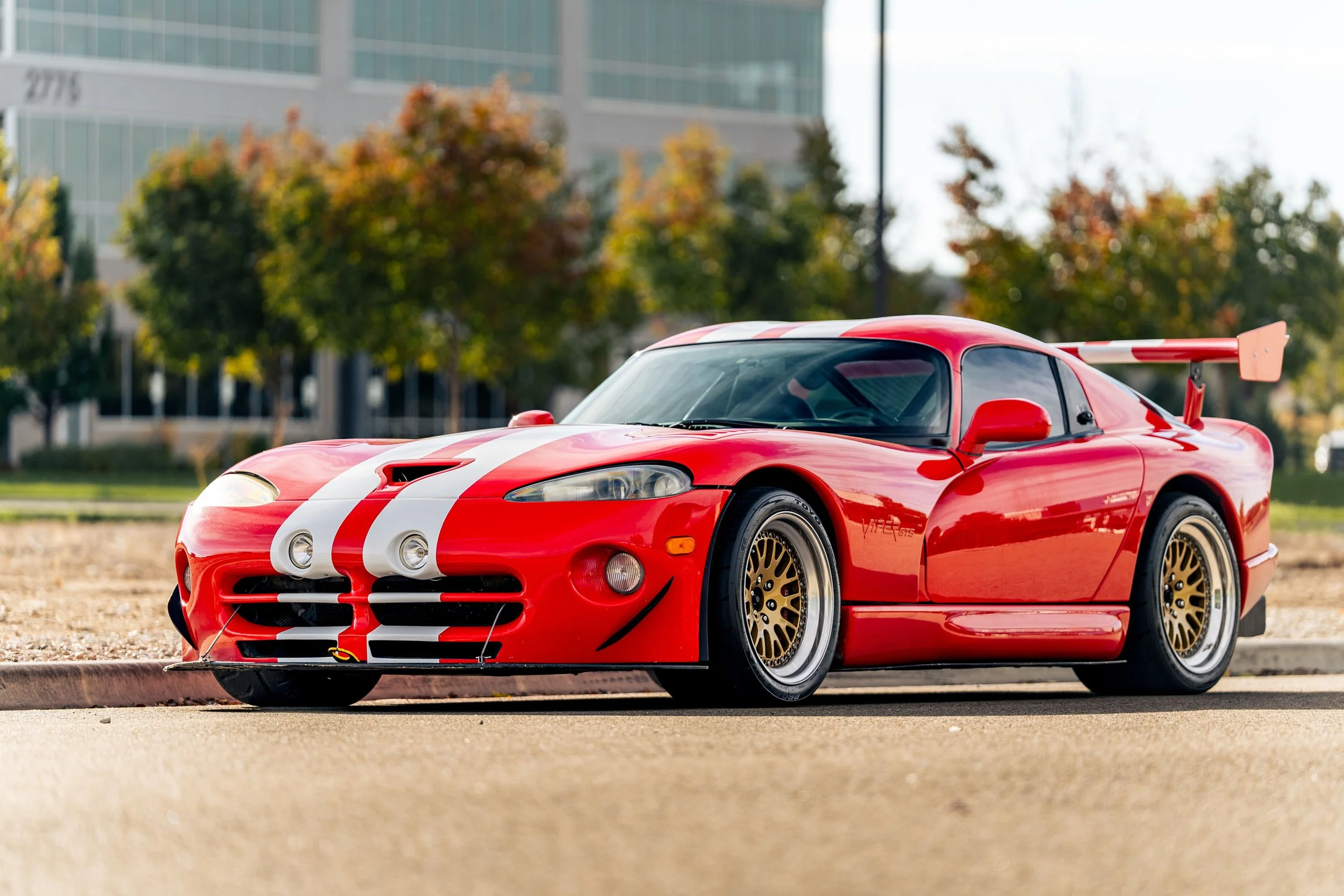 Red race car with white racing stripes parked outdoors on a street or sidewalk with trees and a modern glass building in the background.