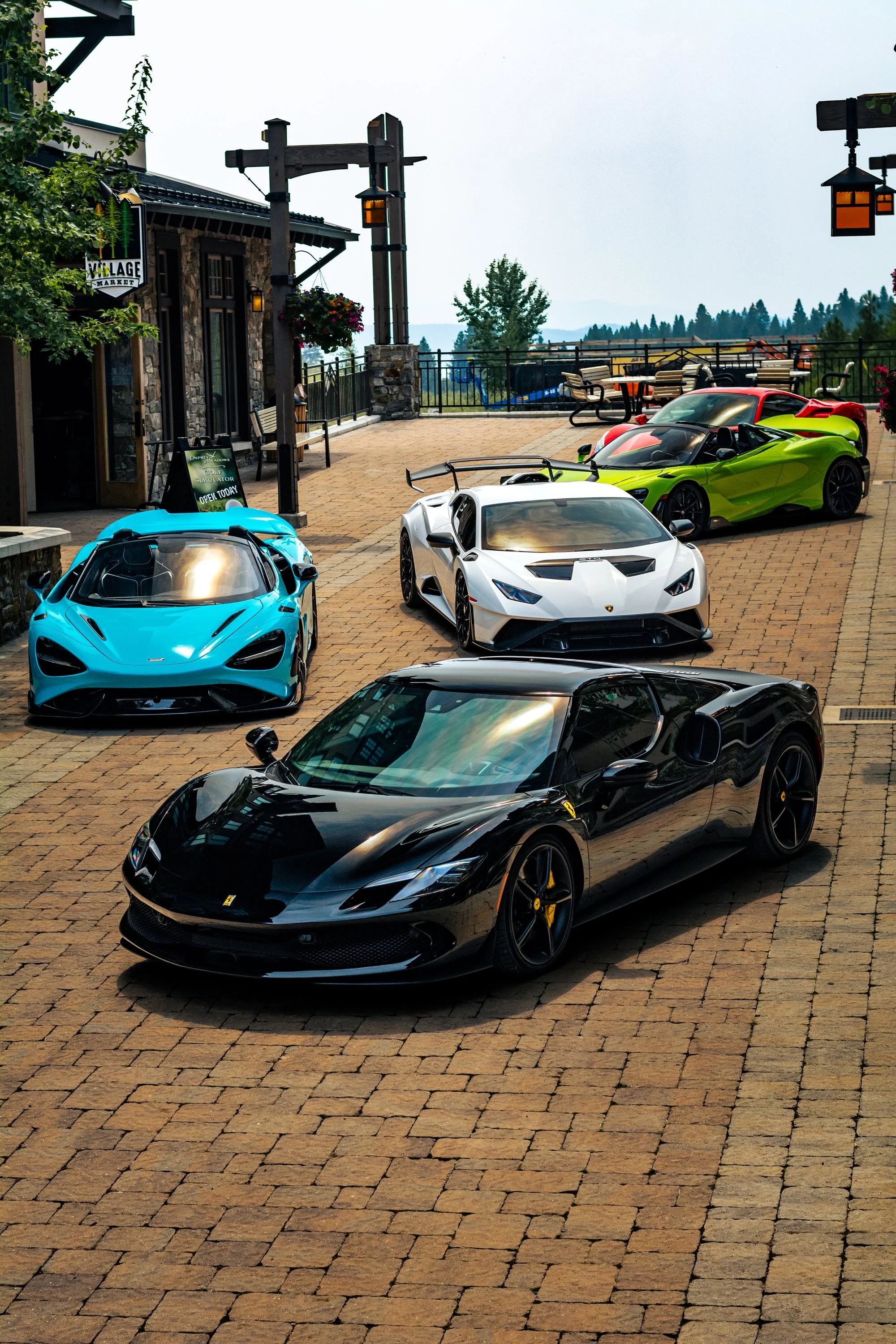 Several luxury sports cars parked on a brick-paved outdoor area with a building to the left, picnic tables in the background, and a scenic landscape beyond. The cars include a black Ferrari, a white Lamborghini, a light blue car, a green car, and a r