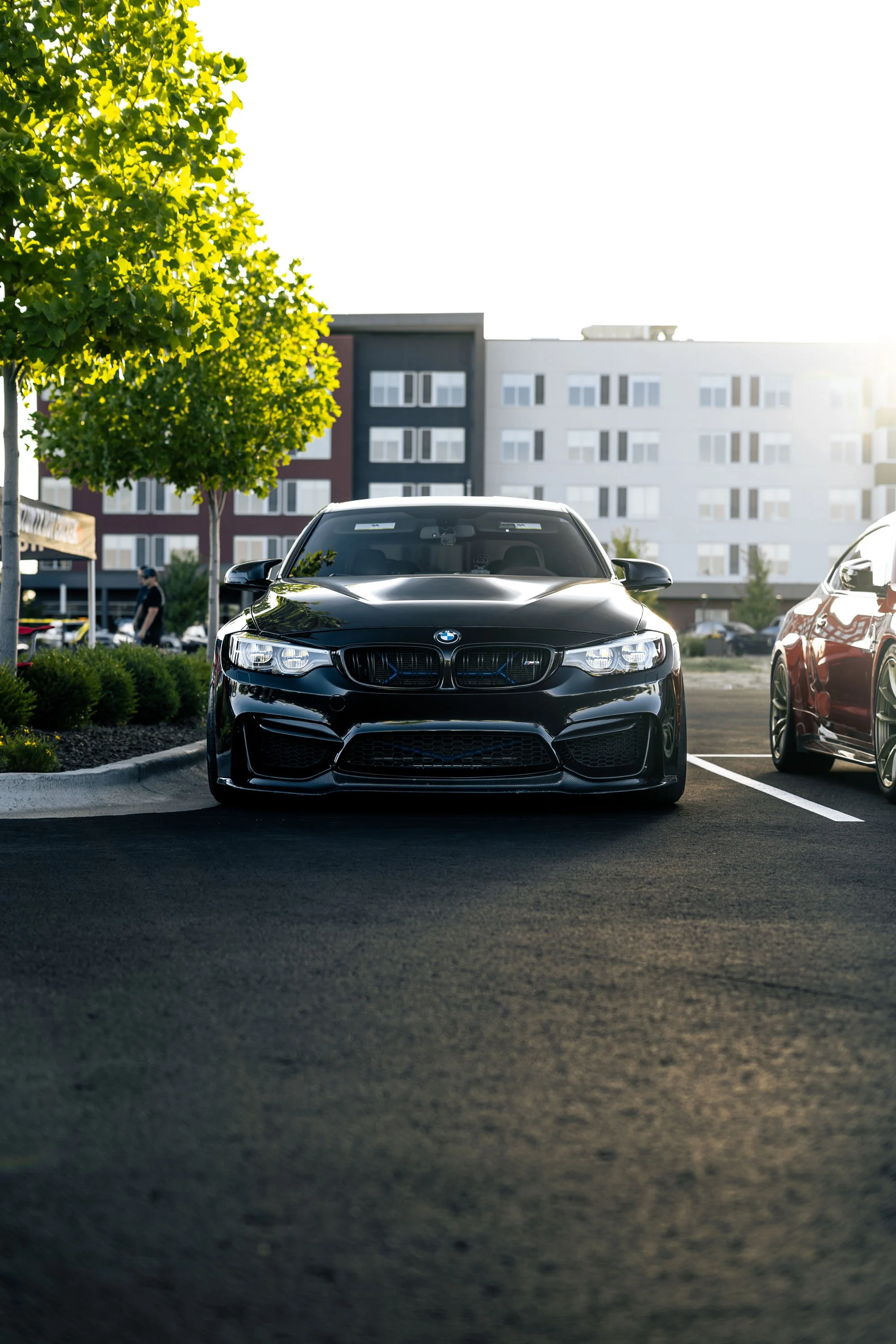 A black BMW M series car parked in a parking lot with buildings and green trees in the background, during daylight.