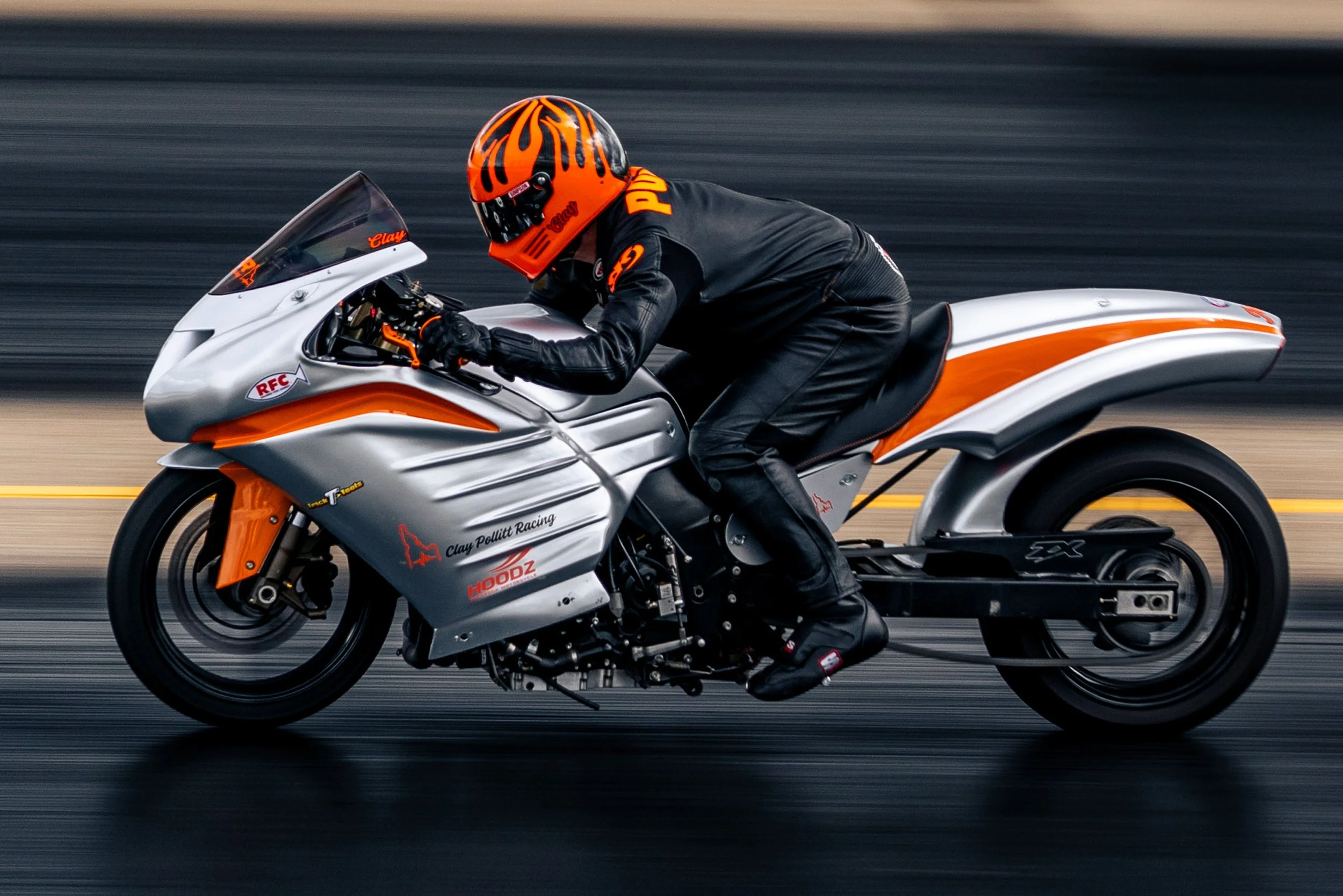 A person wearing a bright orange helmet and black leather racing suit riding a silver and orange motorcycle at high speed on a racetrack.