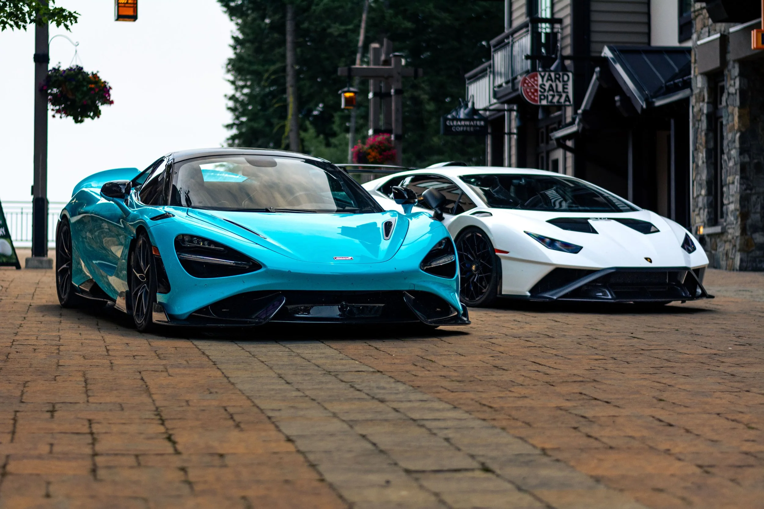 Two sports cars, a blue McLaren and a white Lamborghini, parked on a brick street outside shops with hanging flower baskets and trees in the background.