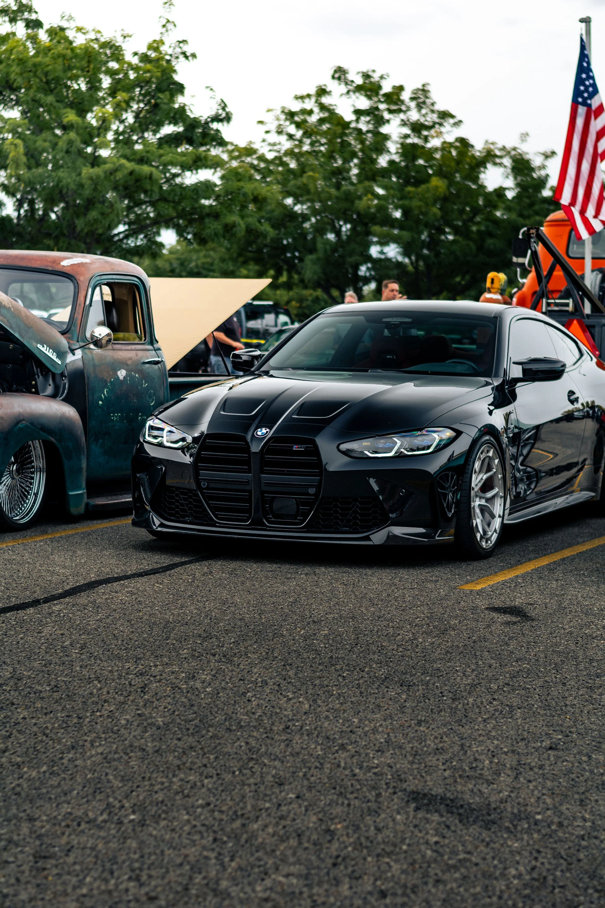 A black BMW car parked next to an old rusty truck, with people walking and a flagpole with an American flag in the background.