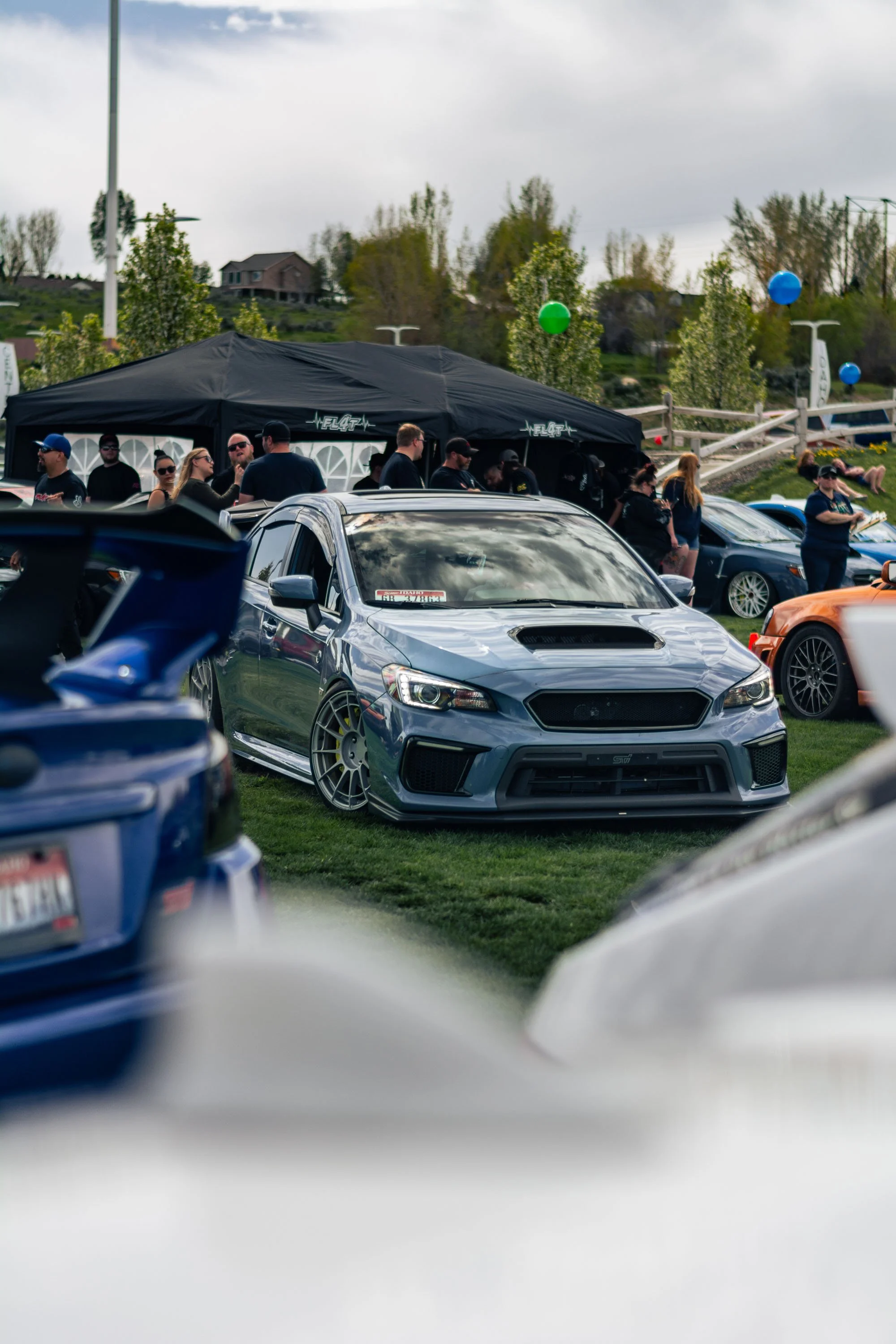 Multiple sports cars parked on grass at an outdoor event, with people gathered around and a black tent in the background, trees, balloons, and a house on a hill under a cloudy sky.
