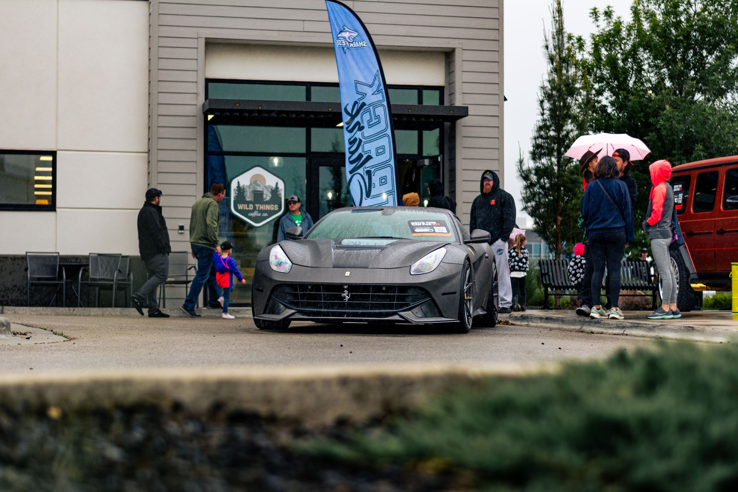 People gathered outside a modern building on a rainy day, with a gray sports car parked in front, a blue flag with 'SHARE THE GE' text, and a mix of children and adults, some holding umbrellas.